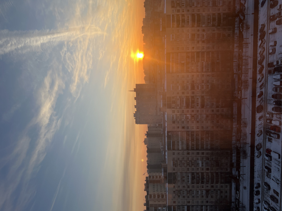 The image depicts a vibrant sunset or sunrise over a dense urban landscape, captured in a portrait orientation but showing a landscape scene if rotated 90 degrees counter-clockwise. The sky dominates the upper and left portions of the image, showcasing a striking gradient of colors from a deep, warm orange and yellow near the horizon where the sun is located, blending into lighter oranges, pale yellows, and finally a soft blue in the upper left corner. Wispy, elongated clouds, possibly contrails, stretch across the blue and lighter orange sections of the sky, catching the light and appearing bright white.
The sun itself is a brilliant, intensely bright yellow orb, appearing just above the horizon line formed by the distant city buildings. Its light creates a strong lens flare effect and casts a warm, golden glow across the entire scene, particularly illuminating the clouds and the upper parts of the buildings.
Below the horizon, stretching across the middle and right side of the image, is a sprawling cityscape composed predominantly of large, multi-story residential buildings. These buildings are mostly concrete or light-colored, with numerous windows and balconies. They appear somewhat uniform in design, typical of a post-Soviet or similar urban development. The closest buildings on the right are quite tall and densely packed, showing many visible individual apartments. Further into the background, more buildings of varying heights can be seen, fading slightly into the atmospheric haze created by the setting or rising sun. One distinct building in the mid-ground, slightly to the left of the sun, has a pointed, almost spire-like roof.
On the ground level, to the right and bottom of the image, there is a large parking area covered in snow, with many cars parked in rows. Adjacent to the buildings, there are bare trees, suggesting winter, and what appears to be a cleared path or road, also lined with snow. The overall impression is one of a cold, clear winter day transitioning into or out of daylight, with the warm hues of the sun providing a strong contrast to the cool blues of the sky and the starkness of the urban environment and snow. No discernible text is visible in the image.