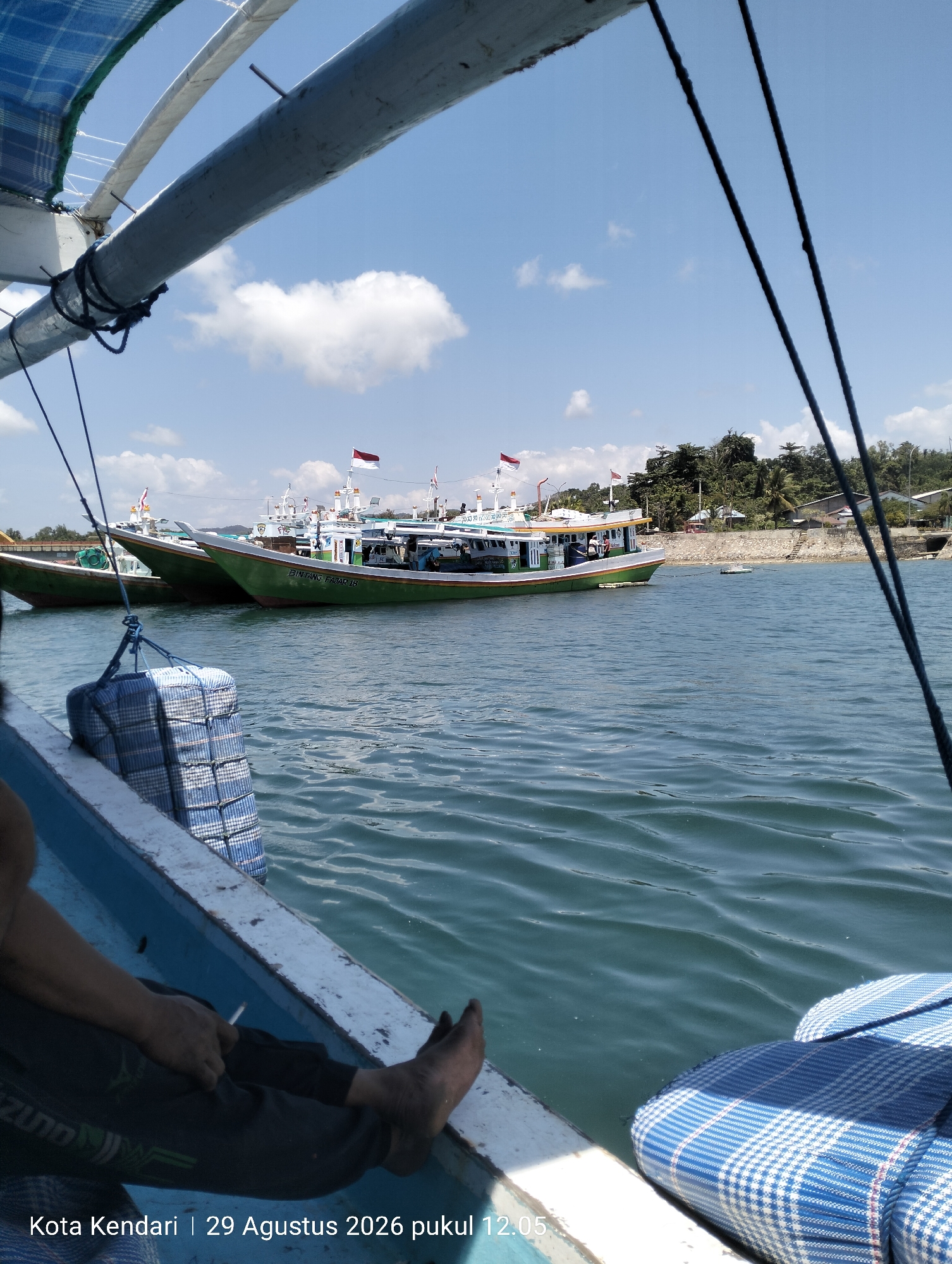 The image, captured on August 29, 2026, at 12:05 PM in Kendari, Indonesia, depicts a serene harbor scene with several fishing boats docked. The perspective is from aboard one of the boats, looking towards a line of larger vessels. In the foreground, a person's legs and feet are visible, suggesting they are relaxing on the boat, possibly smoking a cigarette, with a blue and white checkered cushion nearby. The water is calm with gentle ripples. In the mid-ground, three green and white fishing boats are moored side-by-side, each adorned with flags. The boat furthest to the right has the name "BINTANG FAJAR 13" visible on its hull. Behind the boats, a lush green hillside dotted with houses forms the coastline, under a bright blue sky with scattered white clouds. The overall atmosphere is one of a peaceful, sunlit afternoon in a coastal Indonesian city.
