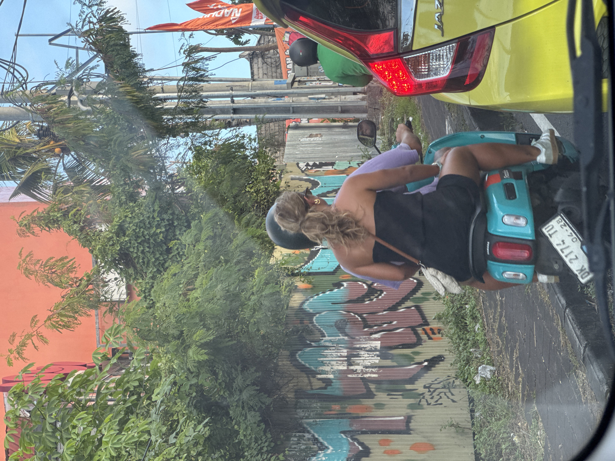 A view from a car window shows a woman on a light blue scooter stopped on the road in Kuta Selatan, Indonesia. The woman is wearing a black top, black shorts, and white sneakers. She has blonde hair and is wearing a black helmet. The scooter has a purple cushion on the seat. A yellow car is directly in front of the scooter. Behind the scooter is a corrugated metal fence with graffiti on it, overgrown with greenery. An orange building and utility poles are visible in the background. Banners hang from the poles. The scooter's license plate reads "DK 2174 TU".