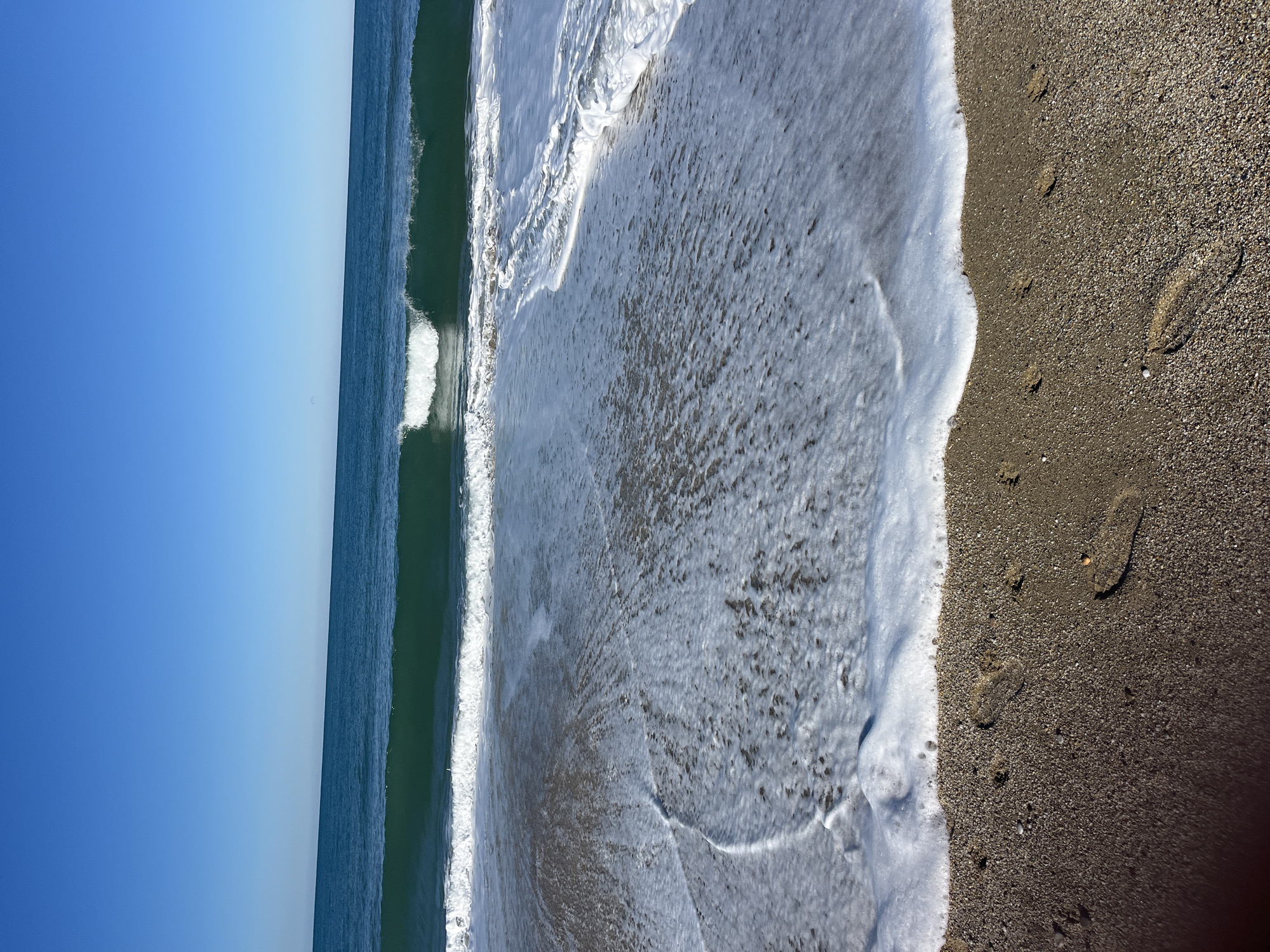 The image shows a beach scene, likely taken at the coastline of Alanya, Turkey. The frame is split vertically: the left side features the sea and sky, with a clear gradient of blue indicating a sunny day. The sea exhibits a dark green color and a wave breaking, marked by white foam. The right side of the image depicts the sand, characterized by a dark brown color. Footprints are visible in the sand, indicating recent human presence. The water meets the sand in a line of white foam, suggesting a receding wave. The overall impression is a peaceful beach scene.