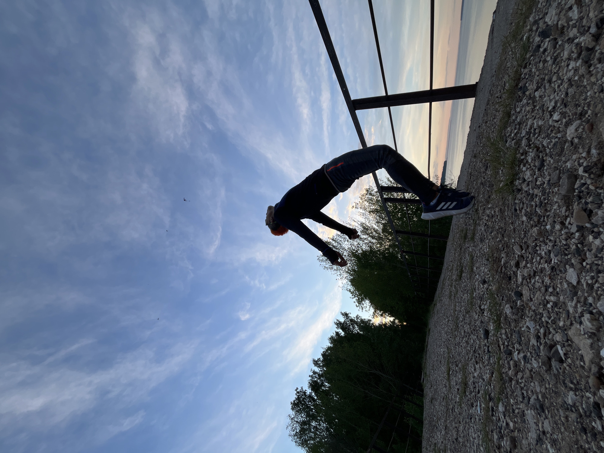 A man in sportswear executes a backflip in front of a metal railing, with a gravel surface beneath him. The background features a line of trees and a distant body of water under a sky with cirrus clouds. The location appears to be a raised platform or overlook, likely in Chaikovsky, Russia, based on the prompt. The image is oriented diagonally, suggesting an unconventional camera angle to capture the dynamic action.