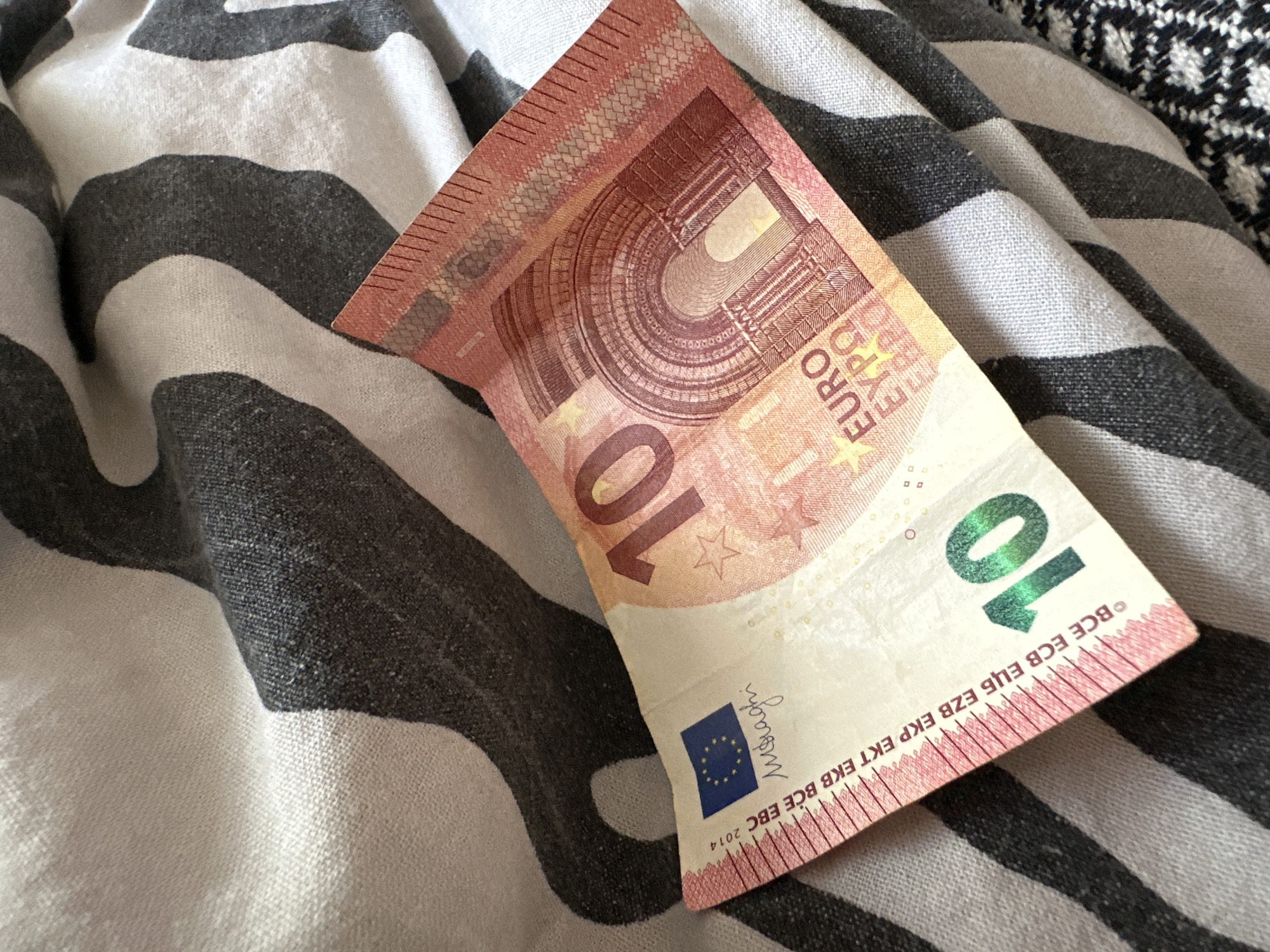 A close-up, top-down photograph captures a 10 Euro banknote resting diagonally on a textured fabric surface. The banknote, primarily in shades of red and orange, displays its front side with a large "10" and architectural designs representing an arch or gateway. Visible text on the note includes 'EURO' in Latin, Greek (ΕΥΡΩ), and Cyrillic (EBPO) scripts, along with the acronyms for the European Central Bank ('BCE ECB ЕЦБ EZB EKP EKT EKB BCE EBC') and the year of issue '2014'. A blue European Union flag symbol is present, accompanied by a cursive signature, which the OCR identifies as 'moraghi'. The numeral '10' also appears in a distinctive green, iridescent effect on the bottom right corner.
The background consists of a soft fabric with wide, wavy stripes in contrasting dark grey/black and light grey/off-white tones, showing folds and texture. A second, finely patterned fabric, possibly houndstooth in black and white, is partially visible in the upper right corner. The scene is static, featuring only the currency and textiles, illuminated by soft, even lighting without any discernible activity or human presence.