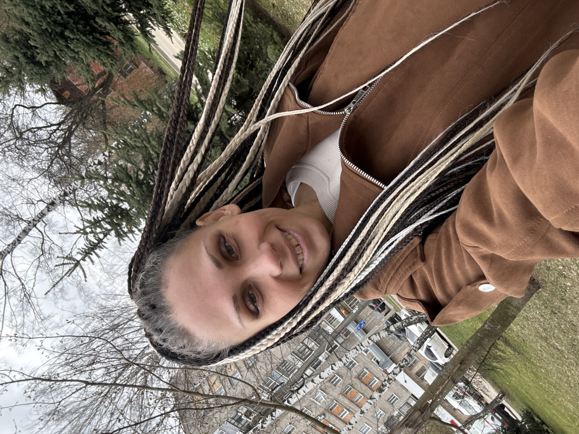 A woman is taking a selfie outdoors in what appears to be a residential area, likely in Rusanovo, Russia, based on the architecture of the apartment buildings visible in the background. The woman has braided hair with black and white extensions. She is wearing a brown jacket and a white shirt underneath. She is smiling at the camera.
The background includes apartment buildings with multiple windows, bare trees, patches of green grass, and parked cars. The sky is overcast.