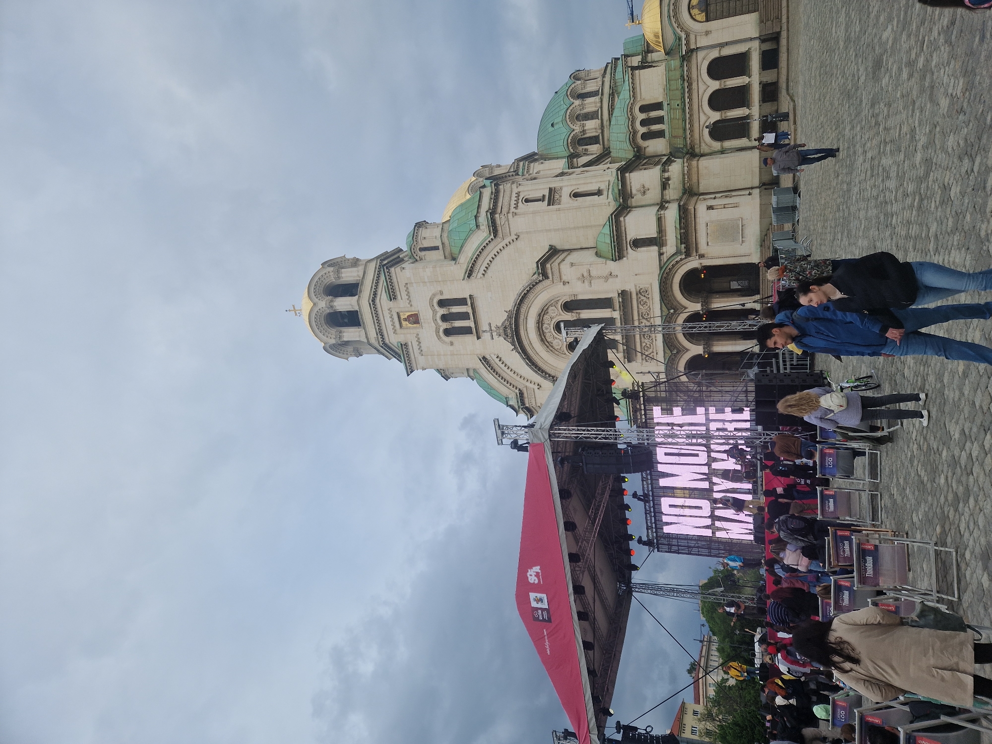 Here is a detailed description of the image:
**Scene:** The image shows a large public gathering in front of a grand cathedral-style building, likely an Orthodox church. The scene is set outdoors, on a cobblestone paved plaza.
**Building:** The building in the background is an imposing structure with a pale stone facade, adorned with multiple green domes and gold accents. It features arched windows, ornate carvings, and classical architectural details.
**Event Setup:** A large stage has been erected in front of the building, indicating an event or performance is taking place. The stage has a large video screen displaying the phrase "NO MORE... MORE". There are rows of chairs set up for the audience, and a red banner or screen is partially visible, possibly as part of the stage design or sponsor branding.
**People:** There are many people present in the scene. Some are sitting in the chairs facing the stage, presumably watching the performance. Others are standing or walking around, including a couple in the foreground.
**Location:** Based on the architectural style of the church, it might be located in a city in Eastern Europe, but the specific location remains unknown.
**Environment:** The sky is overcast with heavy clouds, suggesting a potentially stormy or gloomy weather.