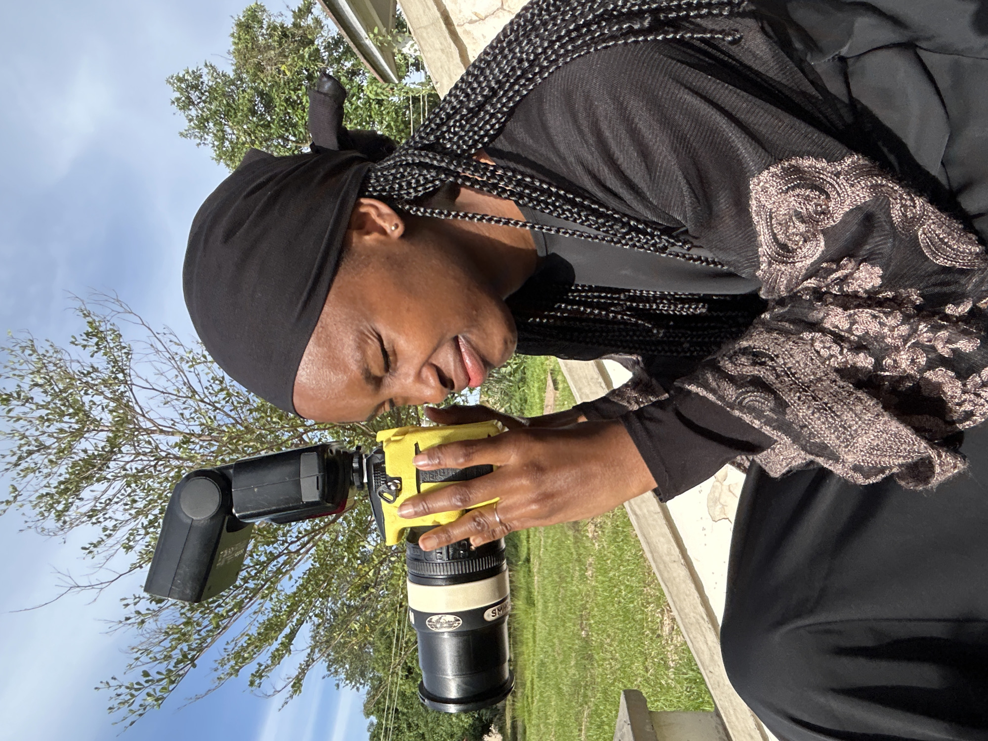 An overhead shot shows a young Black woman with dark skin and black braided hair, wearing a black headscarf and a black abaya with patterned sleeves, holding a yellow camera with a black lens and a flash in her hands. She is looking through the camera's viewfinder and appears to be squinting. The woman is sitting outside, with green grass and a tree in the background. The sky is blue with some clouds. The overall setting suggests an outdoor environment, possibly in a residential area, given the presence of a building facade on the right side of the image. The scene is well-lit, indicating daylight. The location in Vom, Nigeria, is contextual information not directly discernible from the image itself but provided as part of the prompt.