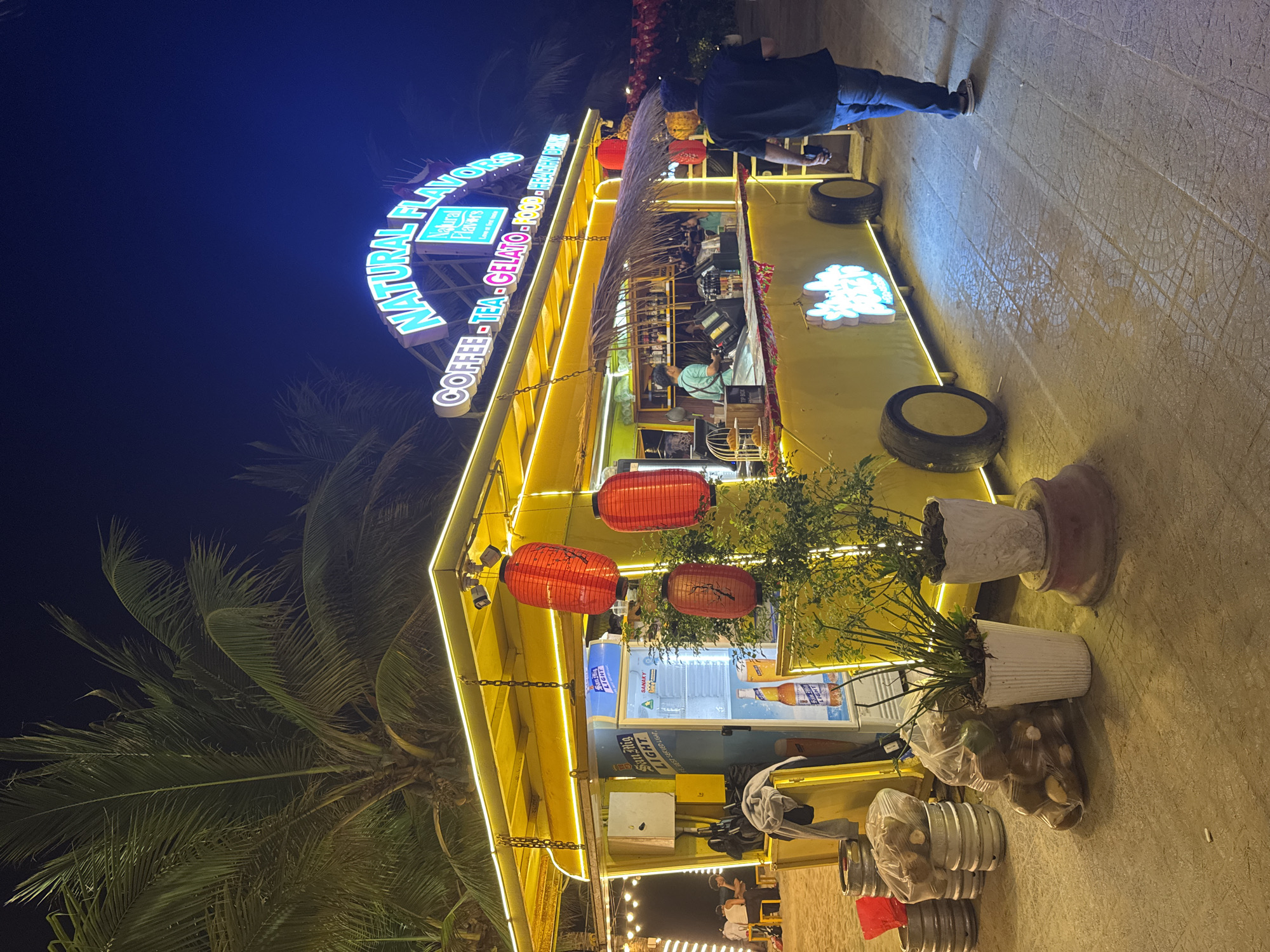 Here is a description of the image.
The photograph shows a brightly lit yellow structure at night. The sign above reads "NATURAL FLAVORS COFFEE-TEA-GELATO-FOOD-HEALTHY DRINK", with the words "Natural Flavors" contained within a leaf logo. Red lanterns hang from the roof. A palm tree is visible on the left.
To the right, a person is standing in front of the building. Plants in pots are positioned near the base of the structure. A refrigerator with the text "San Mig LIGHT" can also be seen.
Based on the visual evidence of the products sold and the general atmosphere, the scene is likely a cafe or a food stall in a tourist area, perhaps on a beachfront. It is located in the city of Đà Nẵng, Vietnam.
