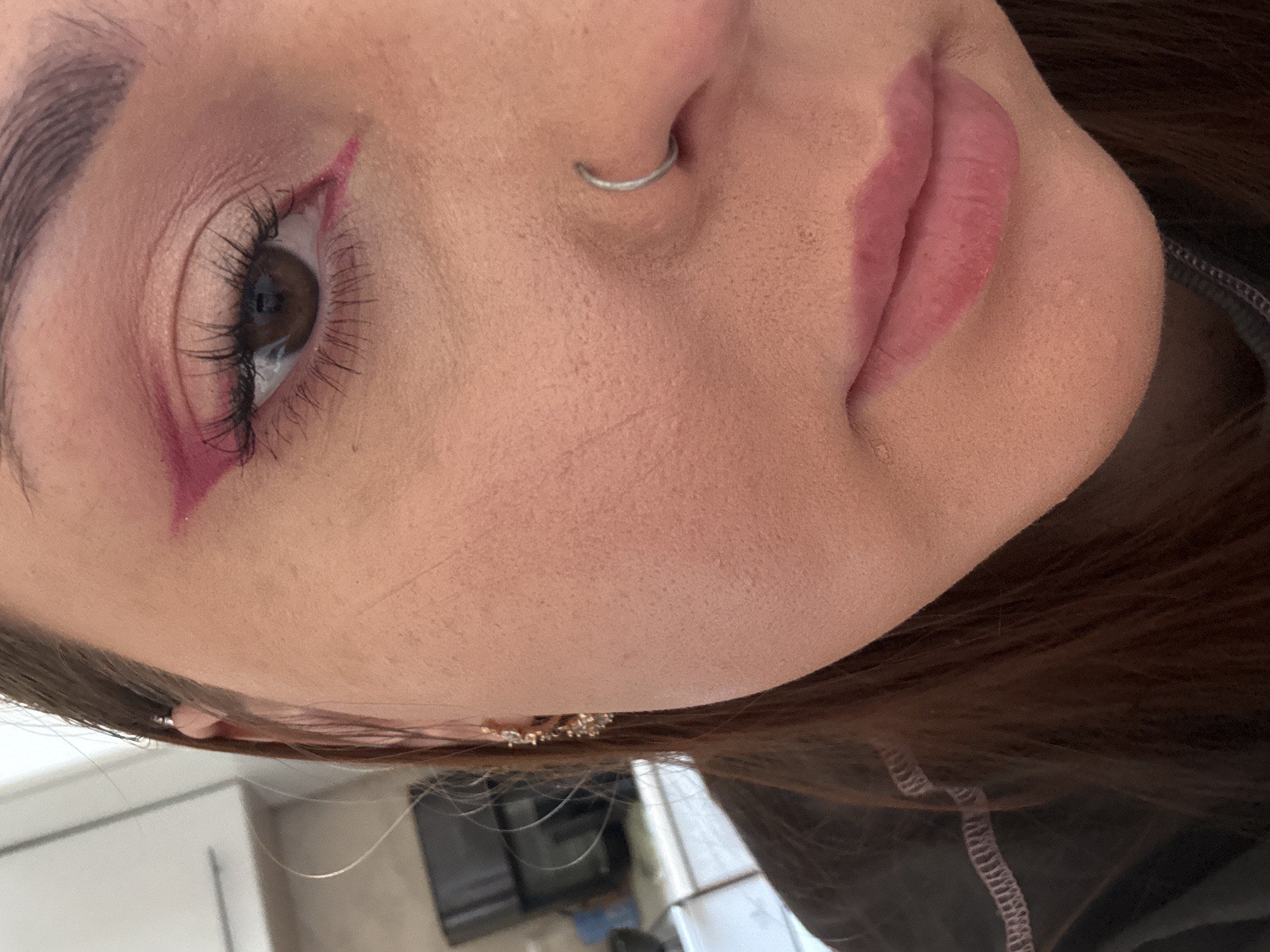 The image is a close-up shot of a person's face, angled to the side. Visible features include an eye with pink eyeliner forming a sharp wing, a nose with a silver ring piercing, and lips with pink lipstick. The person has dark hair. No elements in the photo relate to Izhevsk, Russia. The background is blurred and indistinct, offering no clues about the location.