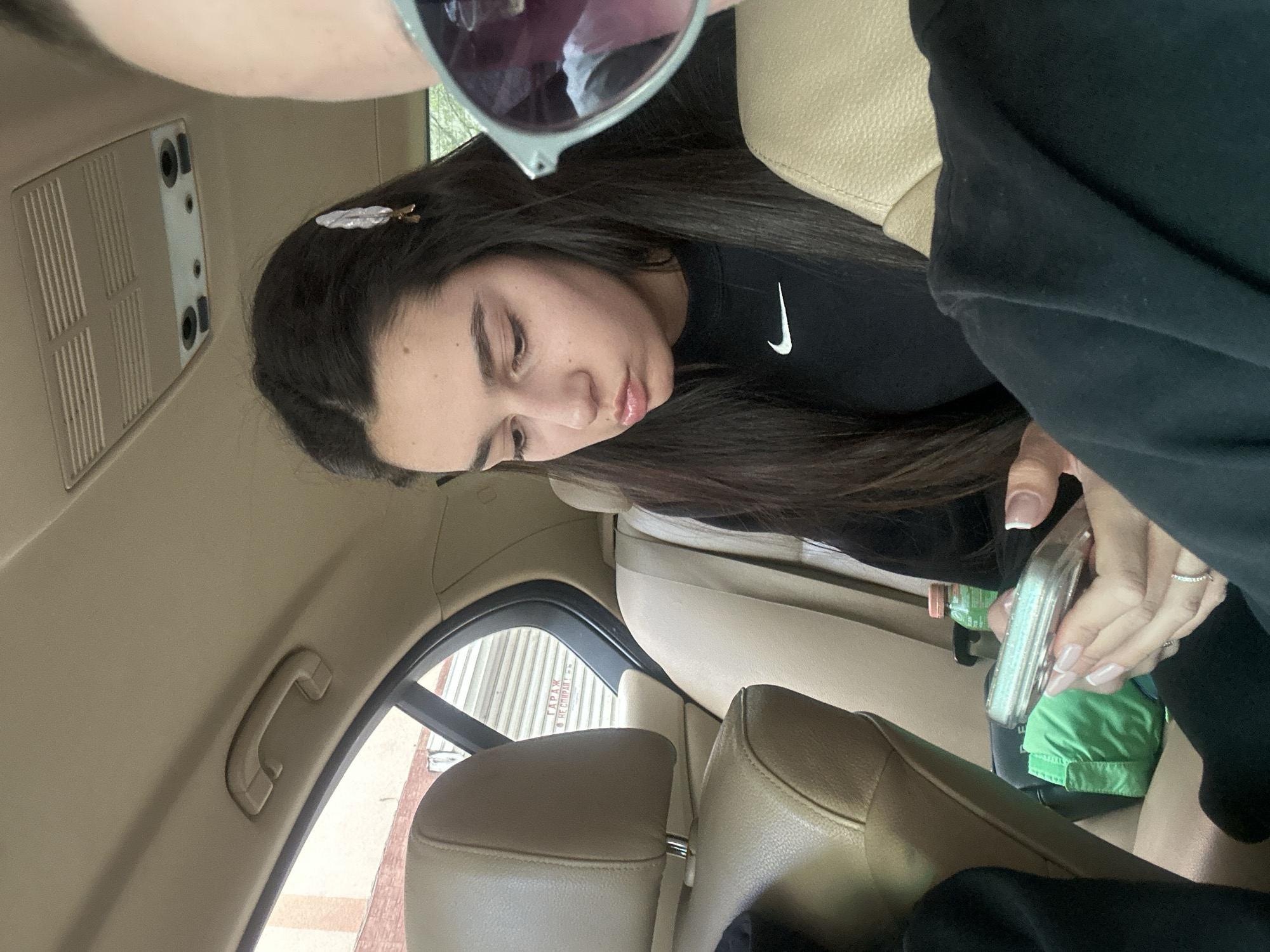 Inside a car, a woman with dark hair and a feather-shaped hair clip is looking down at a cell phone. She is wearing a black Nike sweatshirt and has manicured nails with rings on her fingers. Sunglasses are partially visible above her head. The car's interior is beige leather. The background shows a garage or storage unit exterior with text in Cyrillic, suggesting the location is potentially in Plovdiv, Bulgaria.