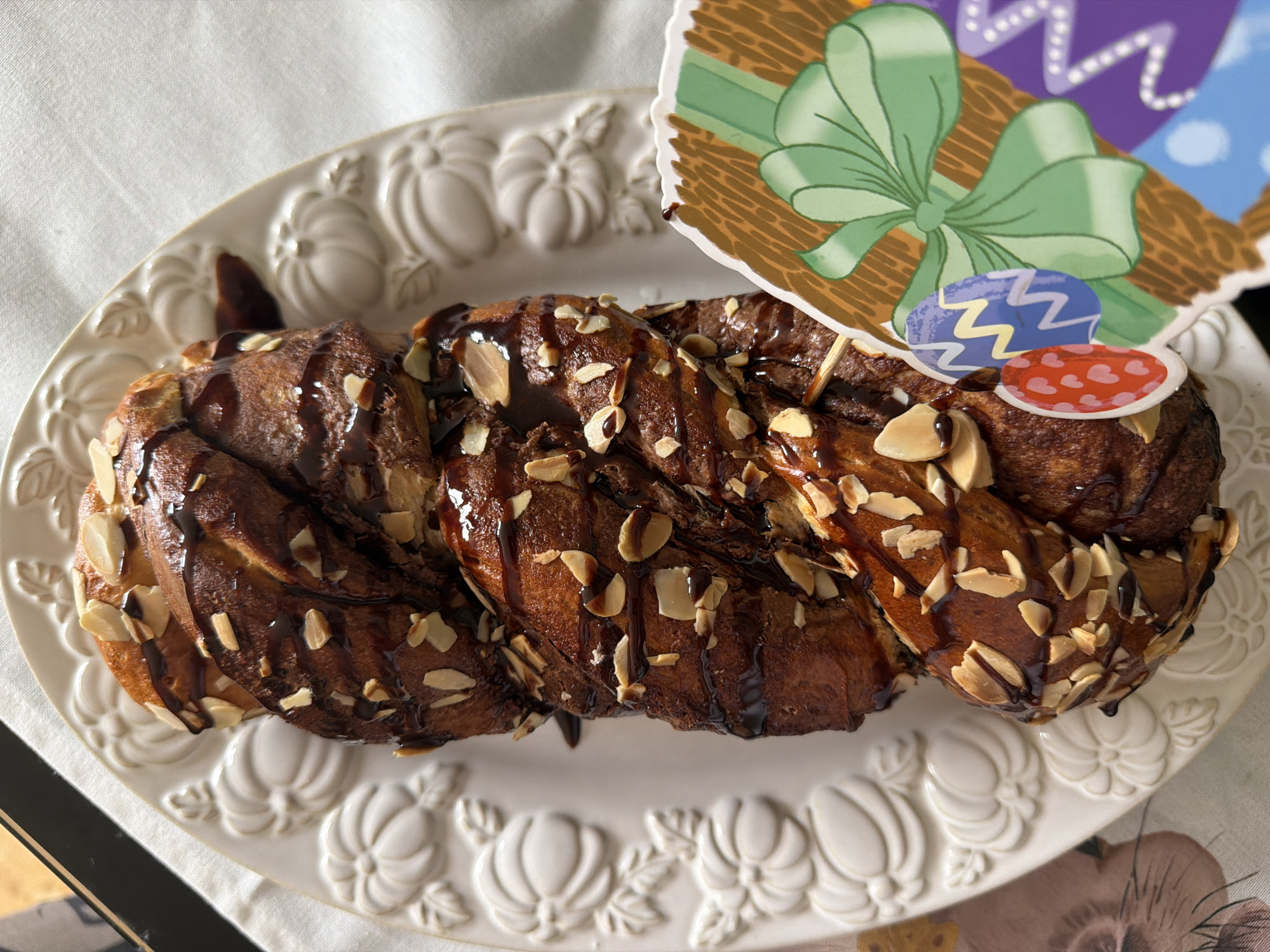 A braided loaf of bread, topped with almonds and chocolate drizzle, sits on an oval, white serving platter embossed with a pumpkin design. An Easter-themed cake topper with a green bow and Easter eggs decorates the loaf. The setting is an indoor environment, likely a home, indicated by the tablecloth beneath the platter.