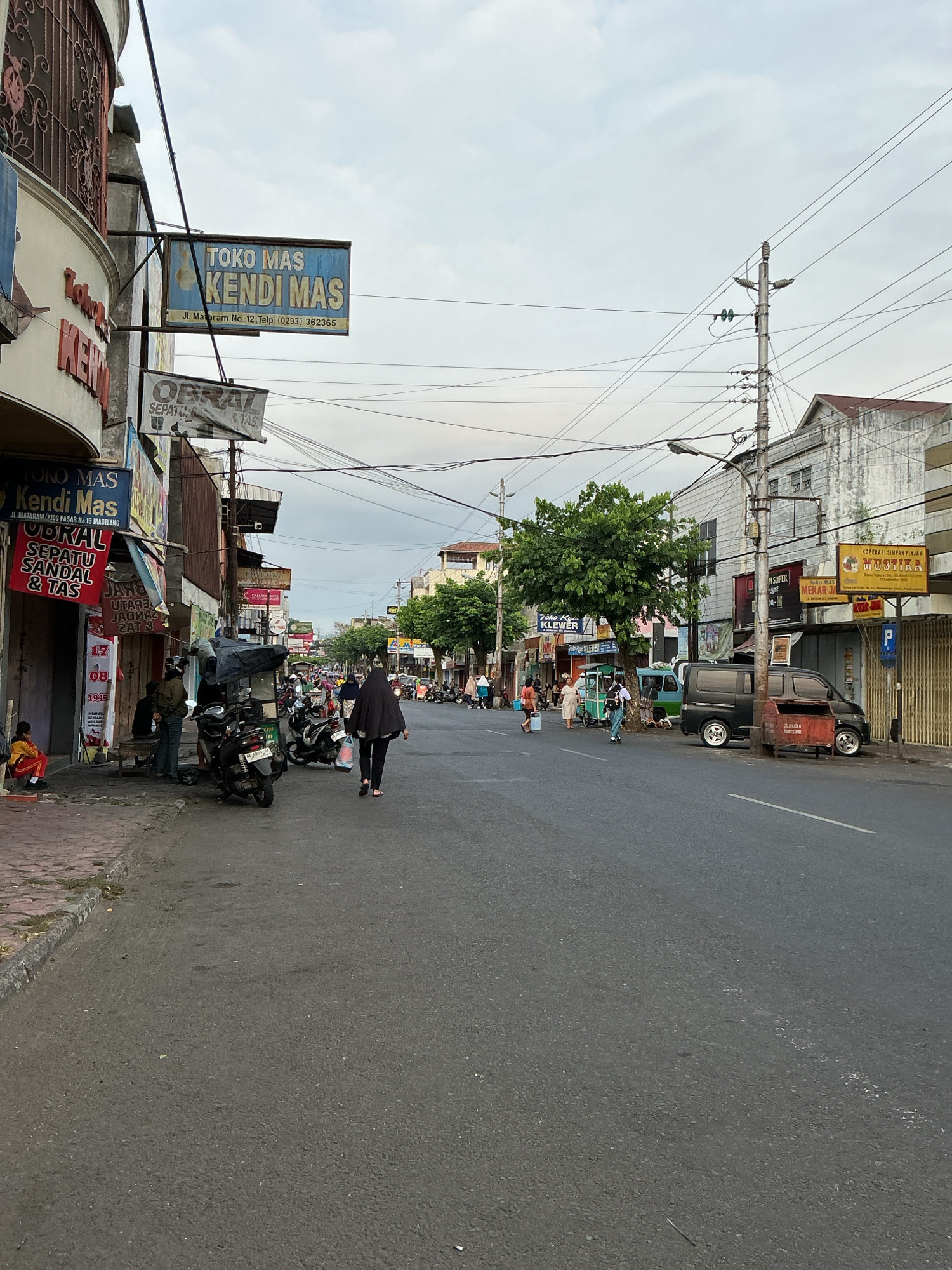 This is an eye-level, medium-shot photograph of a street scene in Magelang, Indonesia. The perspective is from the side of the road, looking down a moderately busy street lined with shops. In the foreground, the asphalt road is visible with a white line marking the lane. To the left, a sidewalk is lined with shops. One shop, "Toko Mas Kendi Mas," has a prominent blue sign with gold lettering. Another sign below it advertises "OBRAL SEPATU SANDAL & TAS" (Sale Shoes Sandals & Bags). Several motorcycles are parked along the sidewalk. A few people are walking on the sidewalk and street. One woman in a black hijab and black clothing is walking away from the camera on the right side of the street.
Further down the street, more shops are visible, including "Toko Mas Mekar Jaya" with a yellow sign. Buildings are multi-story structures, some appearing older and weathered. Power lines crisscross the sky above the street, supported by concrete poles. Trees with green foliage are scattered along the roadside, with one larger tree prominently featured near the center-right of the frame. A black van is parked on the right side of the street. The sky is overcast with diffused light, suggesting it might be early morning or late afternoon. The overall impression is of a typical commercial street in a Southeast Asian city.