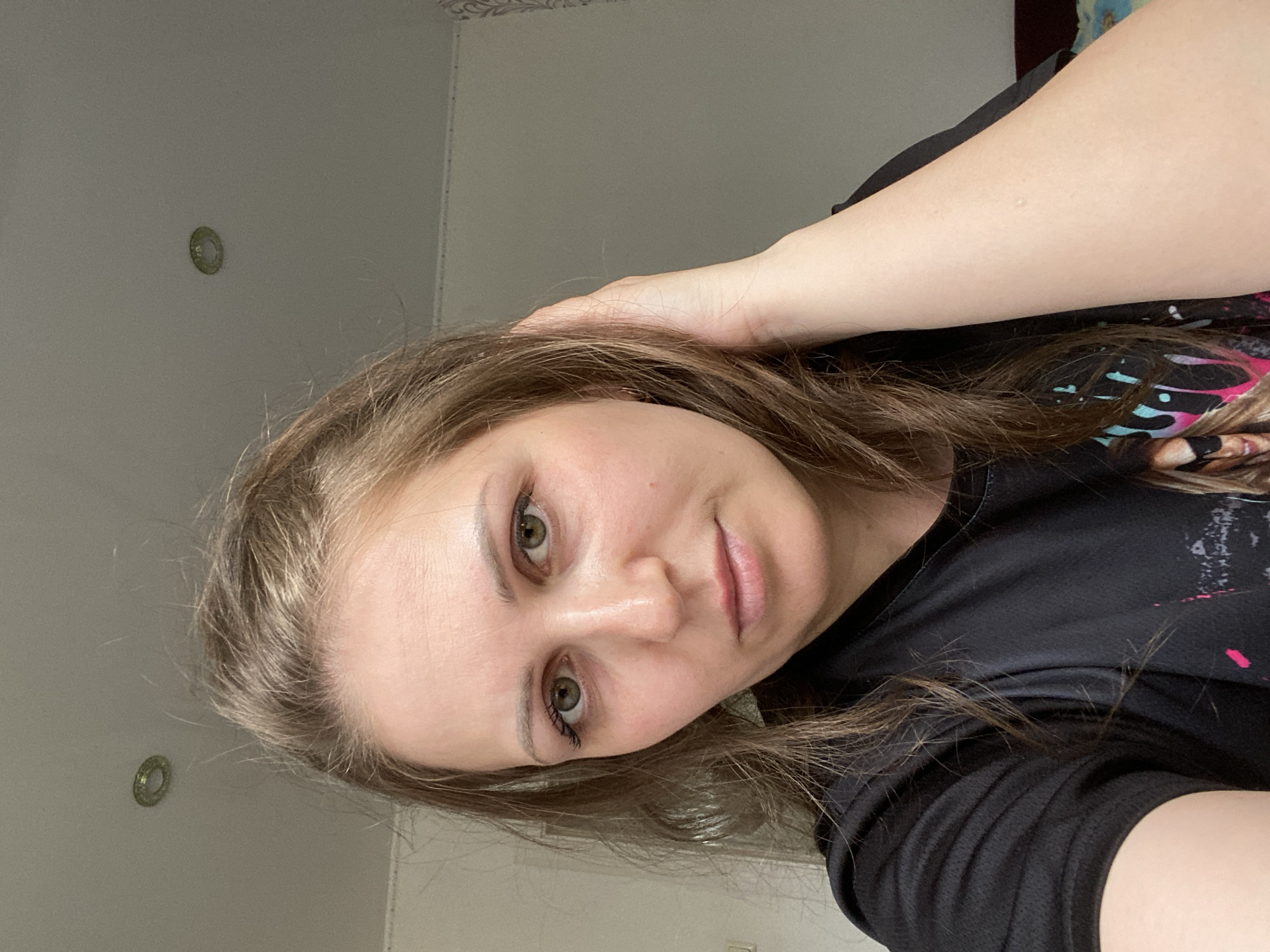 A woman with light brown hair and green eyes is facing the camera. She is wearing a black t-shirt and has her right hand on her head. The background includes a white wall with a circular light fixture. The location is Марковский, Russia.