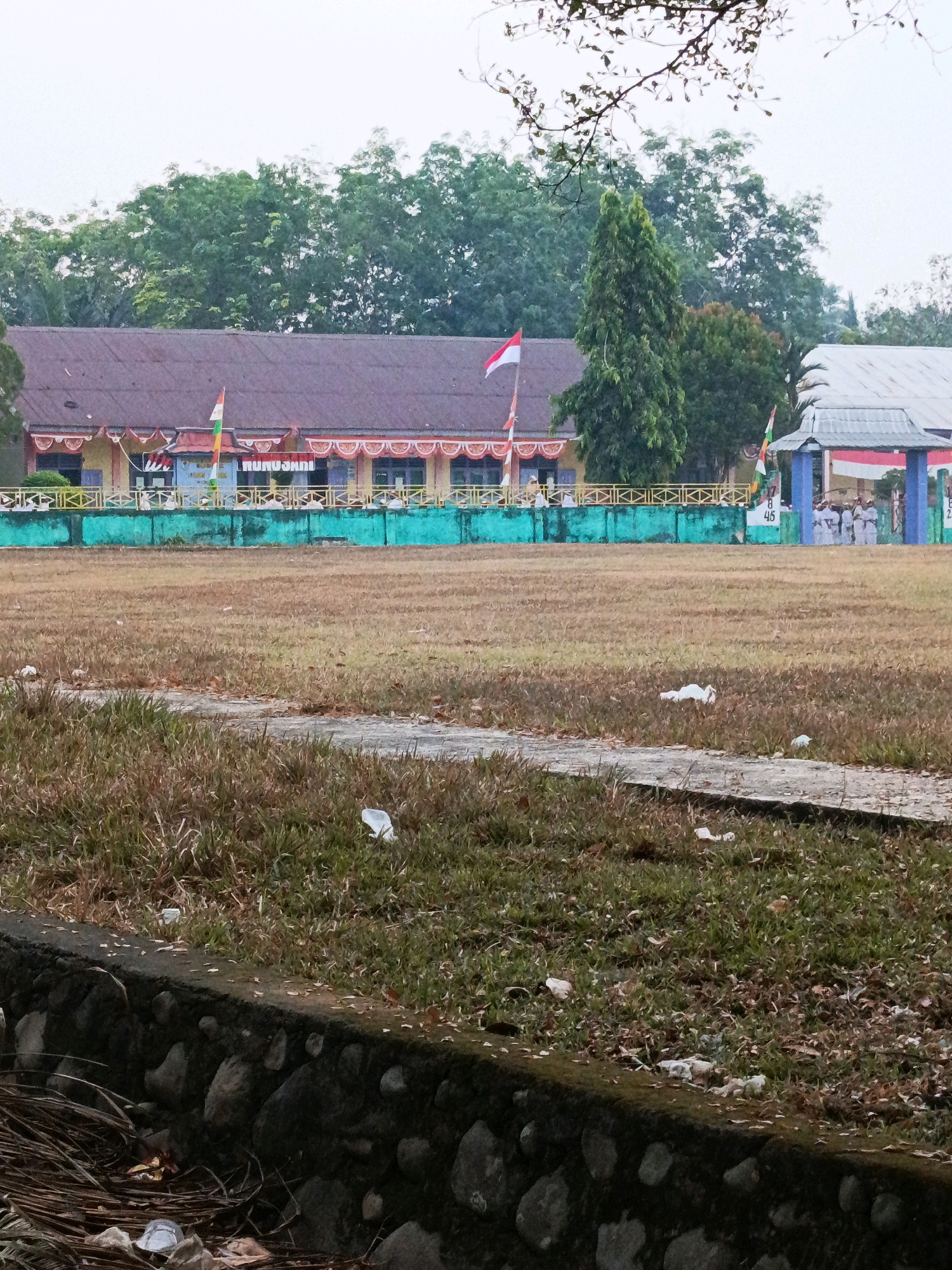 The image depicts an outdoor scene with a building in the background and a dry, grassy field in the foreground. The building appears to be a school or administrative facility with a reddish-brown roof and yellow walls, adorned with decorative banners and flags. In front of the building, there is a green fence and a sheltered area with individuals in uniform. The field is mostly brown and dry, with patches of green grass and scattered debris, including what appears to be plastic waste. A stone-lined drainage ditch runs along the bottom edge of the frame, containing some debris and dried vegetation. The overall environment suggests an overcast day in a semi-urban or rural setting. The specific location is the outdoor field of a school, likely a local government or educational institution in the city of Wonosari, Indonesia.