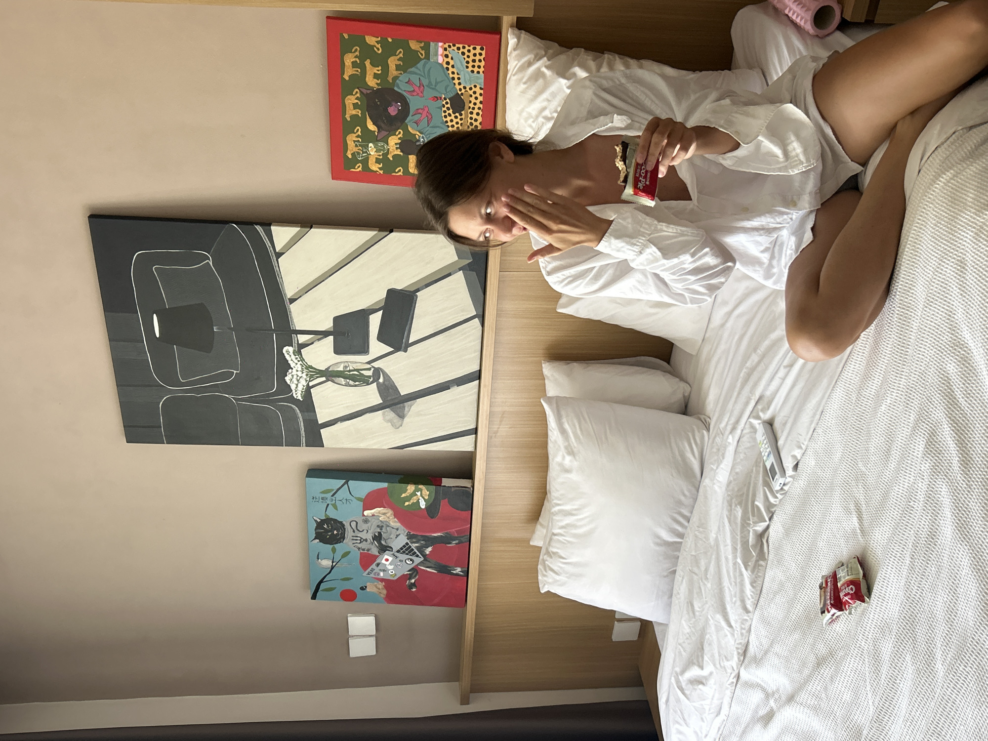 A woman is lying in a bed in a room in North Kuta, Indonesia. She is wearing a white shirt and has a bar of chocolate in her hand. There are three paintings on the wall behind her, one of which is a black and white painting of furniture. There is also a pink foam roller on the bed. The bed has white sheets and pillows.