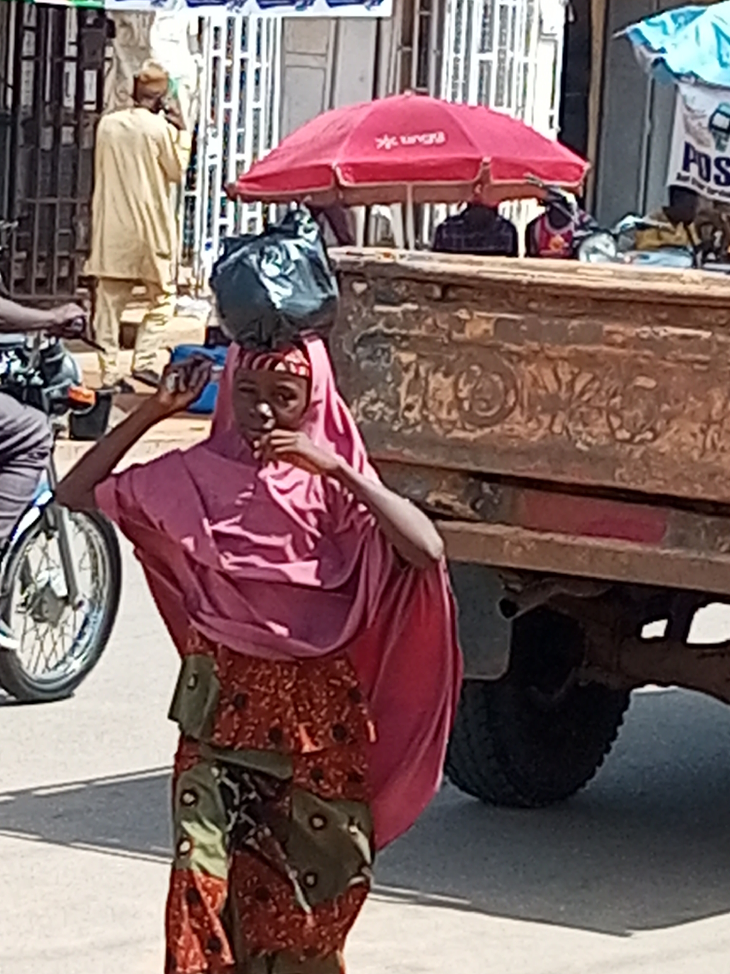 In Gombe, Nigeria, on a sun-drenched street, a young woman carries a large black plastic bag balanced on her head. She is wearing a pink hijab and a patterned dress with green sashes. The woman is walking beside a parked truck. The truck's side shows weathered, textured metal, and its large rear tire is visible. In the background, a man in a light-colored tunic speaks on a phone near a white gate. A red umbrella, advertising "Ungu," shades a market stall. The environment suggests a bustling marketplace with goods displayed under awnings, including a blue tarp with the word "POS" visible. The ground is paved, and shadows stretch across the scene, indicating the time of day.