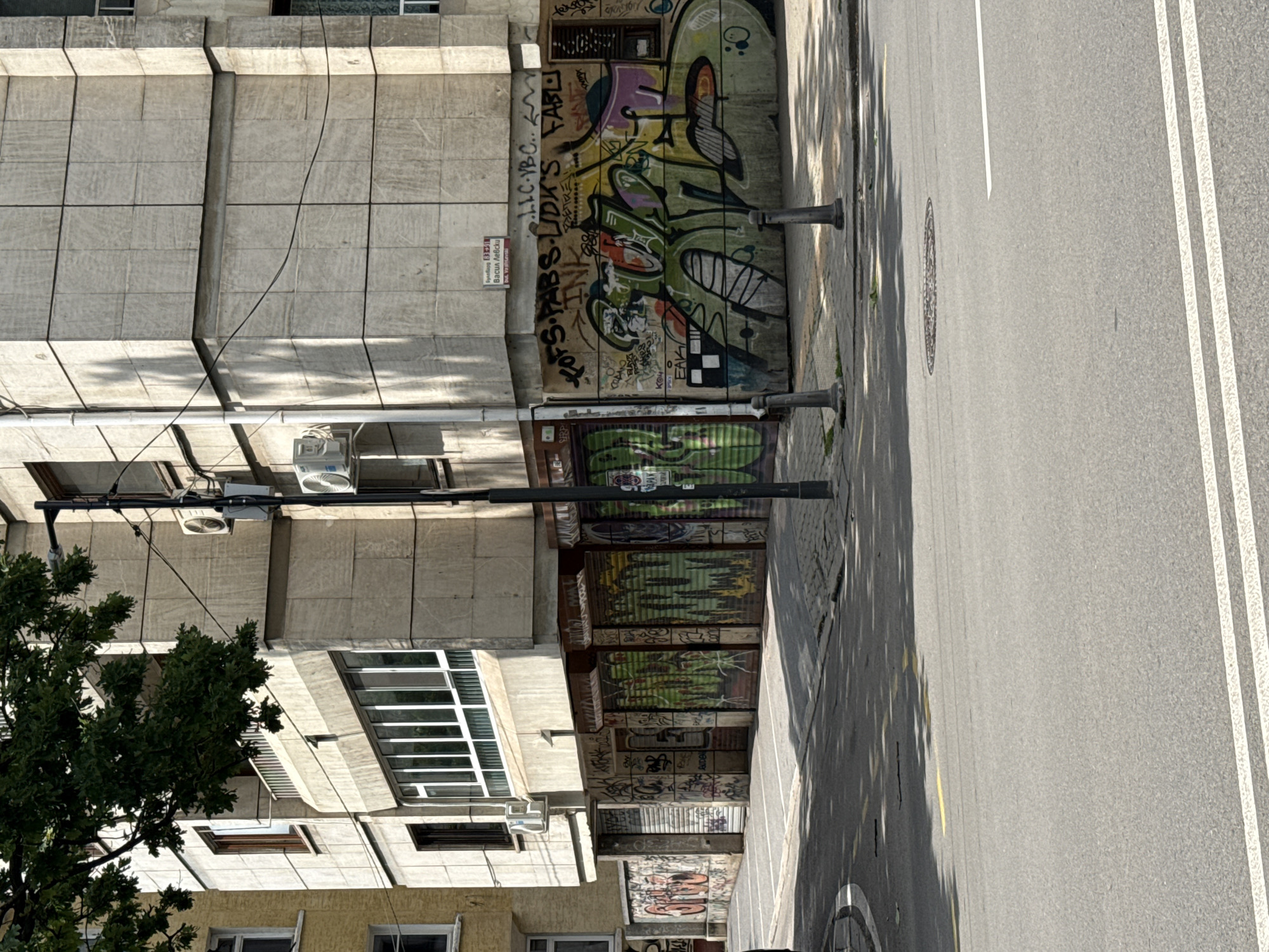 An urban streetscape, rotated 90 degrees clockwise, depicts a sunny daytime scene in what is confirmed to be Sofia, Bulgaria. On the left, a multi-story concrete building features large rectangular panels and various windows, including prominent gridded ones on lower floors. Several air conditioning units and electrical wires are visible on its facade. The lower sections of this building and adjacent structures are extensively covered in vibrant and monochrome graffiti, including on metal roll-down shutters of storefronts. A smaller, yellow building is partially seen at the bottom.
To the right, an asphalt road runs parallel to the buildings, marked with clear white double-lane lines and a single white line. A circular manhole cover is embedded in the street. A paved sidewalk separates the road from the buildings, featuring a dark street pole or lamp post. A green tree on the far left casts long, distinct shadows across the sidewalk and part of the road, suggesting a bright day with the sun at a lower angle.
Visible text includes a red street sign identifying the location as "Васил Левски" (Vasil Levski) and "bul Vasil Levski". Graffiti elements include "LLC.VBC...", "FOFS PABS LIDK'S FABL", "INK", "EAK", and "Ko".