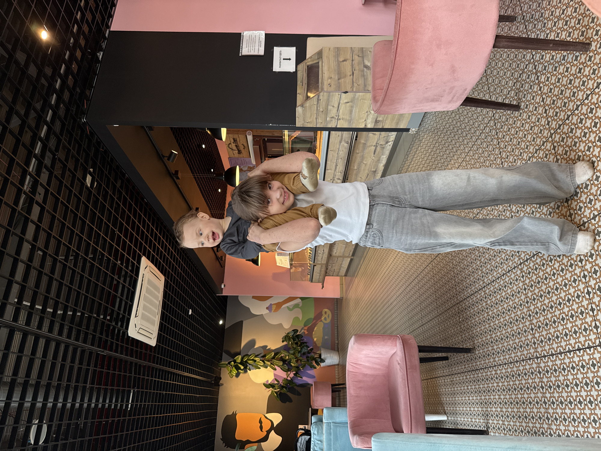 Two children are in a café. One child is carrying the other on their shoulders. The carried child has a brown teddy bear. The cafe has pink and blue chairs and a patterned tile floor. The wall has a mural painted on it. The cafe is possibly in Rusanovo, Russia.