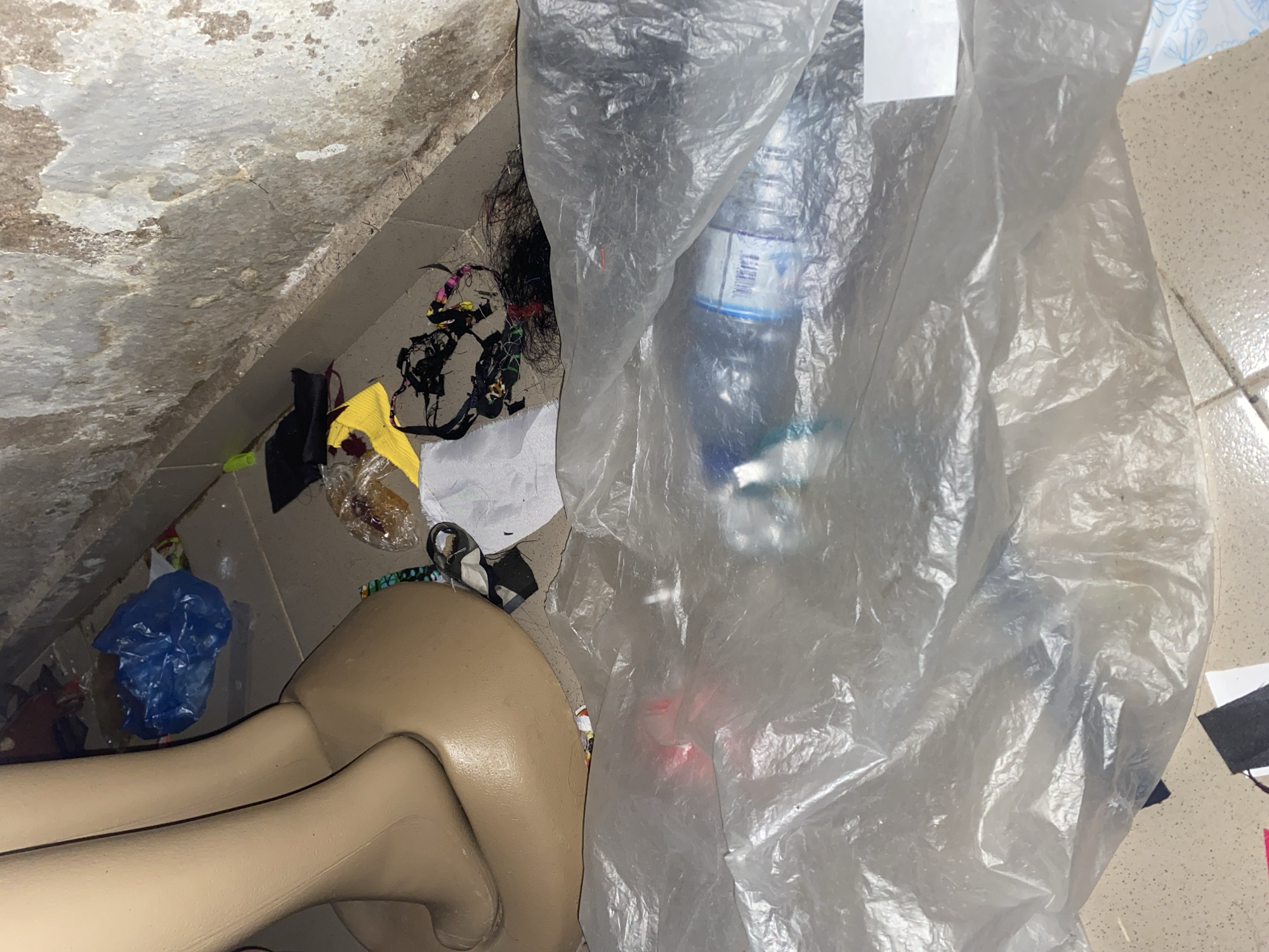The image depicts a cluttered environment with various discarded items. In the foreground, a large, translucent plastic bag obscures a significant portion of the scene, containing a plastic water bottle. Behind this, a beige mannequin leg and base are visible, resting on a tiled floor. Scattered around the mannequin and on the floor are pieces of fabric, plastic bags, and what appears to be debris, including a crumpled blue plastic bag and some dark, stringy material possibly representing hair or thread. A textured, weathered wall forms the background. The scene is dimly lit, contributing to a somewhat unkempt and abandoned appearance. The location is identified as Jauro Yinu, Nigeria.
