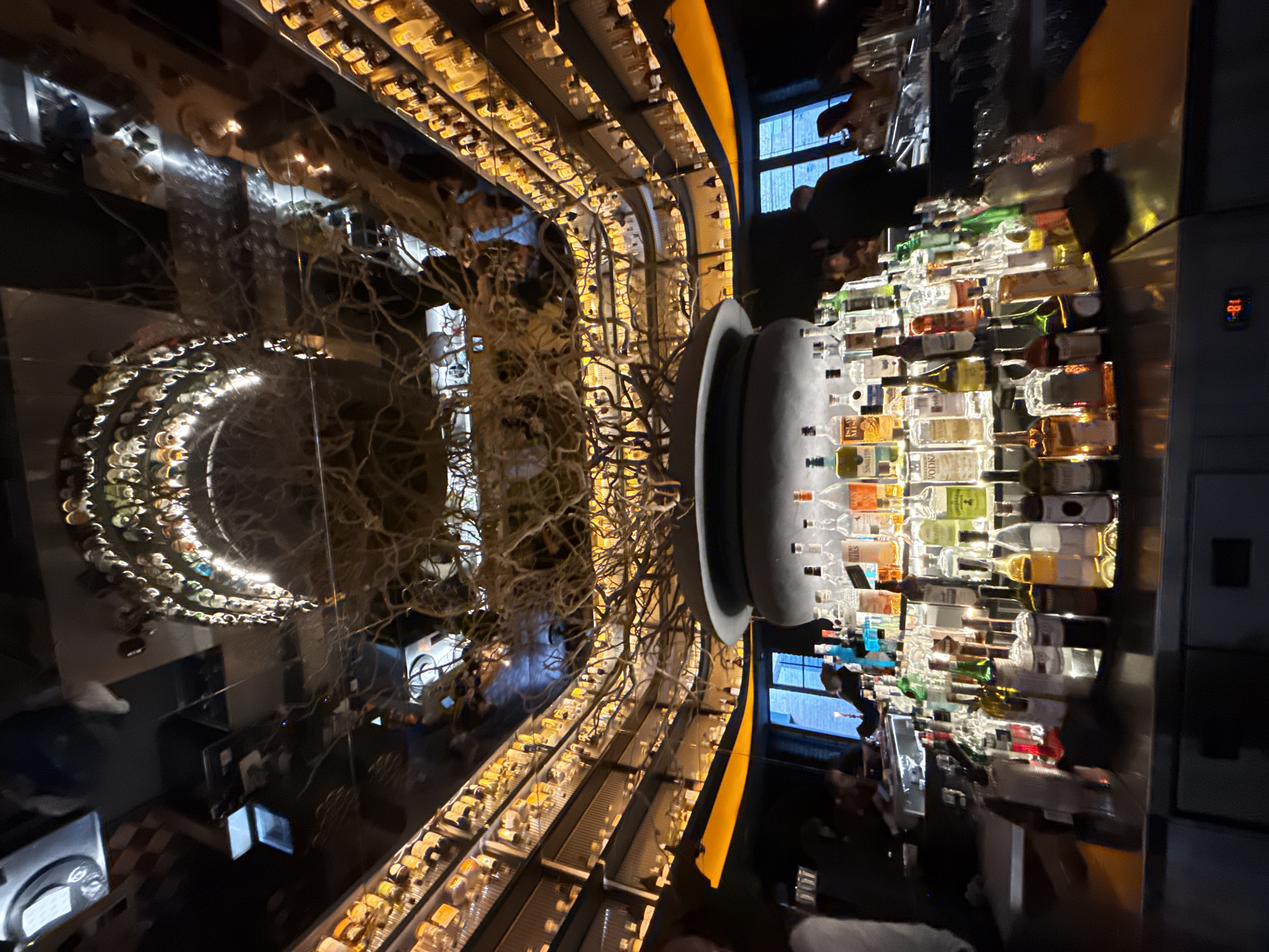 The image depicts the interior of a dimly lit, upscale bar or restaurant. The scene is dominated by rows of liquor bottles displayed on shelves that curve upwards and line the walls, illuminated by warm orange lighting. A large, dark, cauldron-like structure sits prominently in the center foreground, adorned with dry, bare branches that extend upwards, creating an artistic centerpiece. The ceiling is mirrored, reflecting the bottle-lined shelves and the central branch structure, adding depth and a sense of grandeur. In the foreground, a bar counter is laden with various bottles of alcohol, some clear and others colored, with an assortment of glasses. Several people are visible in the background, appearing to be patrons seated at tables or standing near the bar, their faces indistinct due to the low lighting and distance. The overall ambiance suggests a sophisticated and possibly theatrical environment. This scene is located in the city of Vinnytsia, Ukraine.