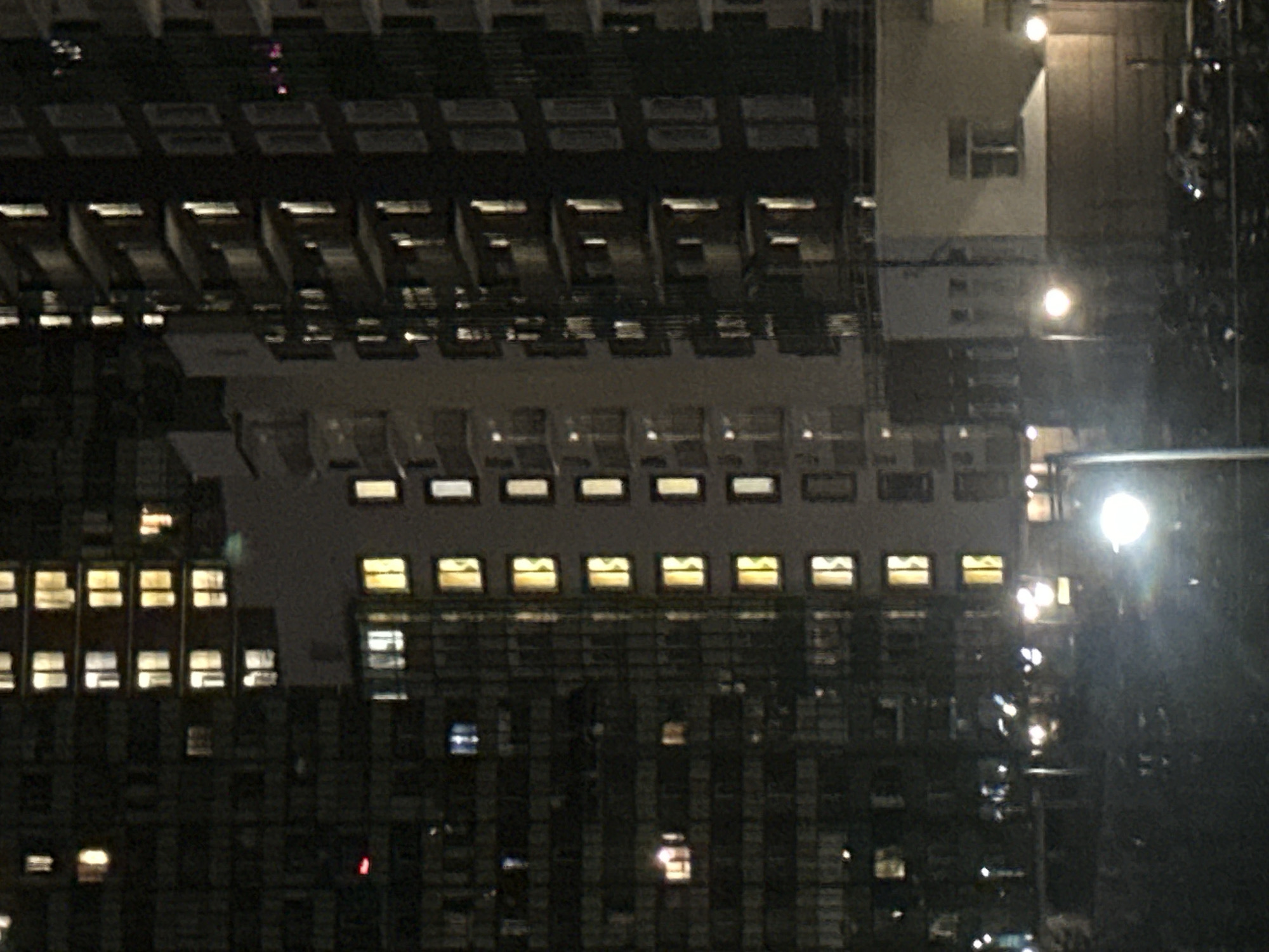 The image depicts a night scene of apartment buildings in Sinyavino, Russia. The buildings are illuminated by numerous lit windows, creating a pattern of light against the dark facades. Some buildings have balconies with railings. In the foreground, streetlights and car headlights create bright, blurry light sources. The ground level appears to be a dark, possibly paved area with some vehicles visible. The overall atmosphere is urban and nocturnal.