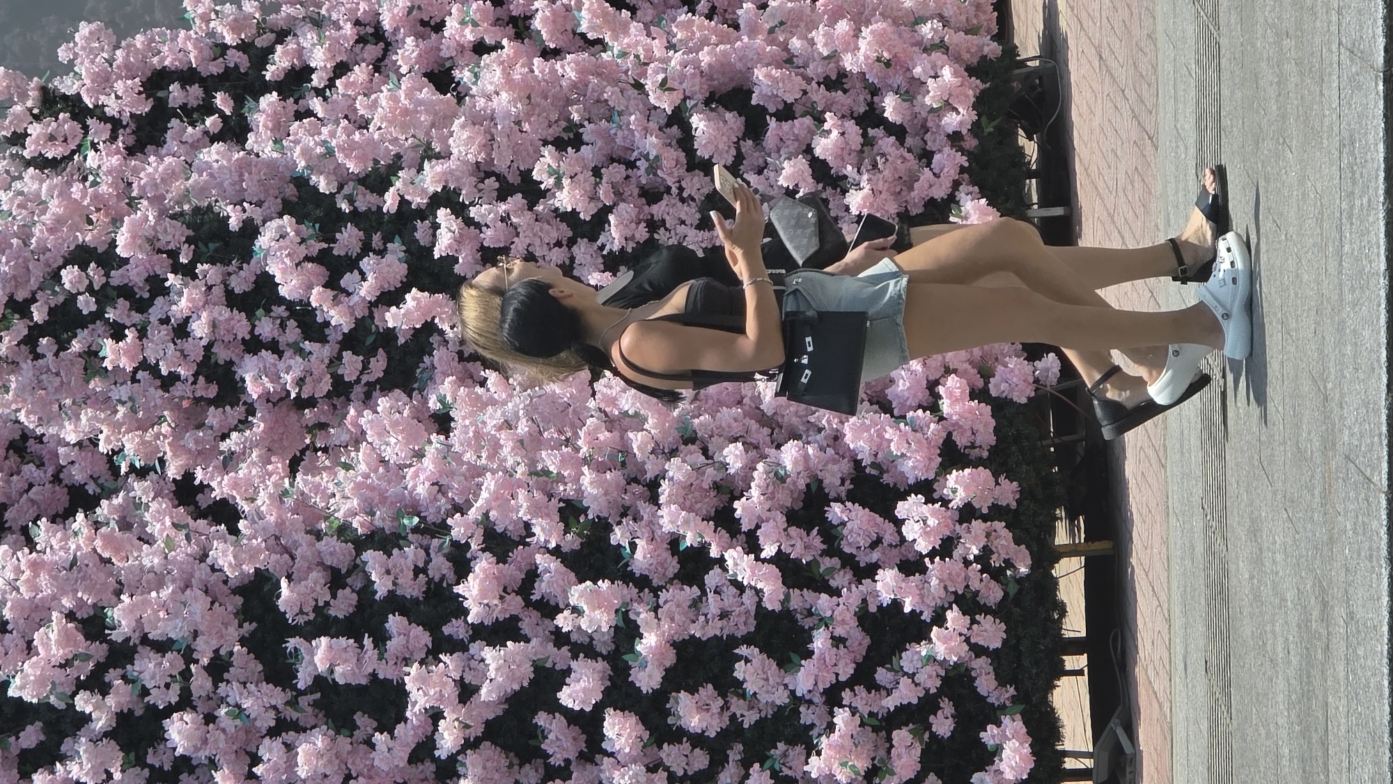 Two women stand in front of a large display of pink artificial cherry blossoms in Nha Trang, Vietnam. The woman on the left has blonde hair, sunglasses, and wears a black tank top and denim shorts. She holds a phone in her hands. The woman on the right has dark hair pulled back and also wears a black tank top. A black purse hangs from her shoulder. Both women are standing on a sidewalk, and the woman on the right wears white Crocs.