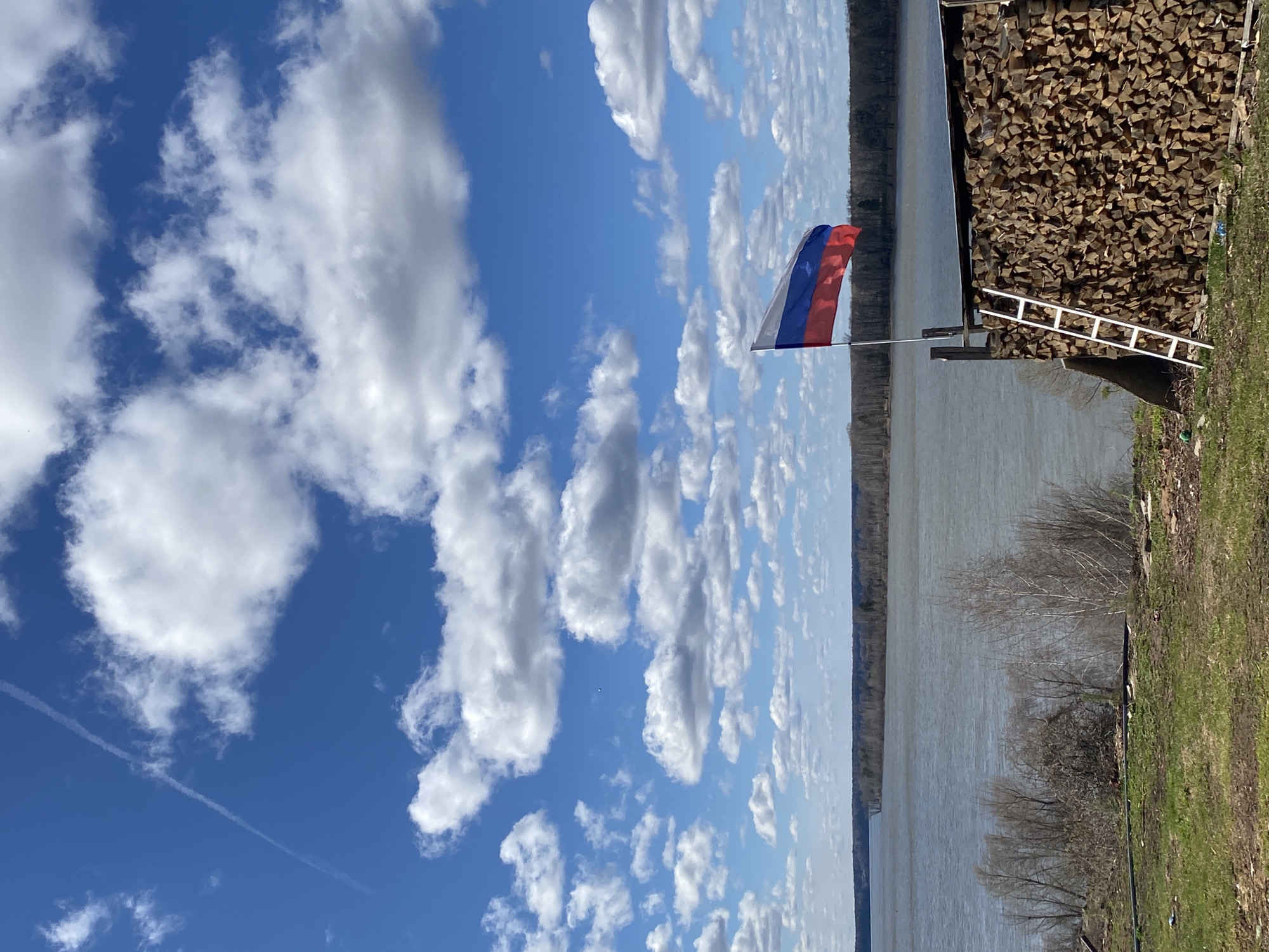 The image shows a landscape in Ольховка, Russia. A Russian flag is prominently displayed against a blue sky filled with white, puffy clouds. Adjacent to the flag is a large stack of cut firewood. To the right is a white ladder propped against the woodpile. Beyond the firewood is a body of water, likely a river or lake, with the opposite bank visible in the distance. Near the body of water, there are trees with bare branches and patches of green grass. A contrail stretches across the left side of the sky.