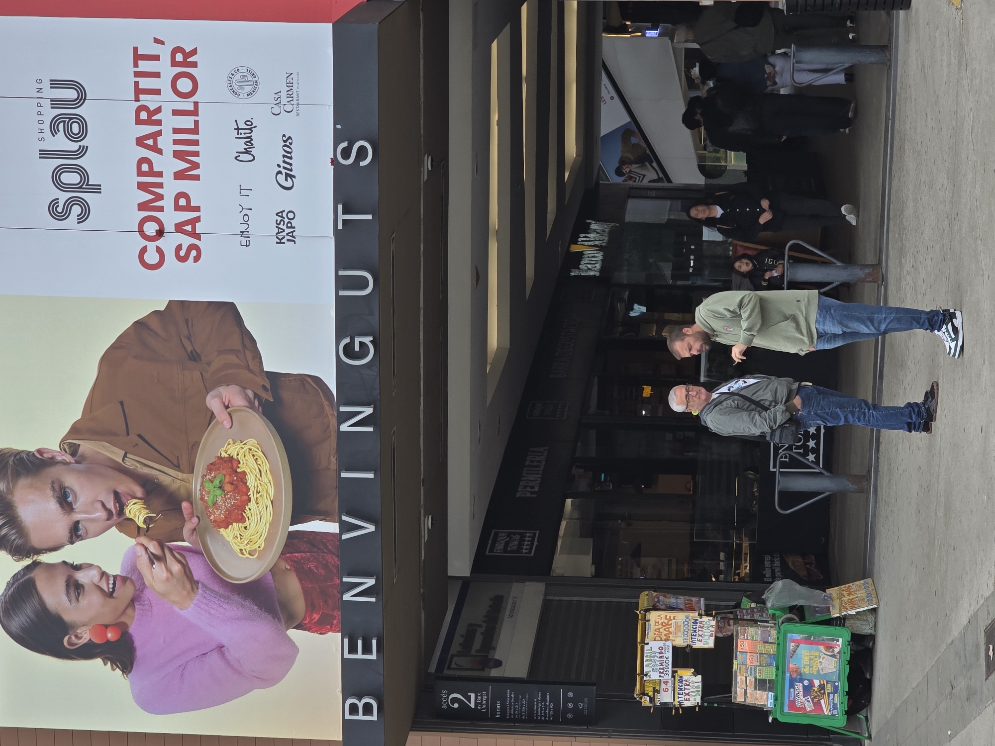 The image captures a street scene in el Prat de Llobregat, Spain. A large advertisement for the Splau shopping center dominates the view, featuring a couple sharing a plate of spaghetti. Below the ad, the entrance sign "BENVINGUTS GUTS" is visible.
Several people are present on the street. Two men stand in the foreground: one wearing a green shirt and jeans, the other wearing a grey jacket. In the background, several people are visible, including a woman seated on a bench.
A newsstand displays various lottery tickets and magazines. The storefronts behind the people feature signs for "PERNILERIA" and "ENRIC TOMÁS." A sign indicates the address "2 av Baix Llobregat" along with opening hours.