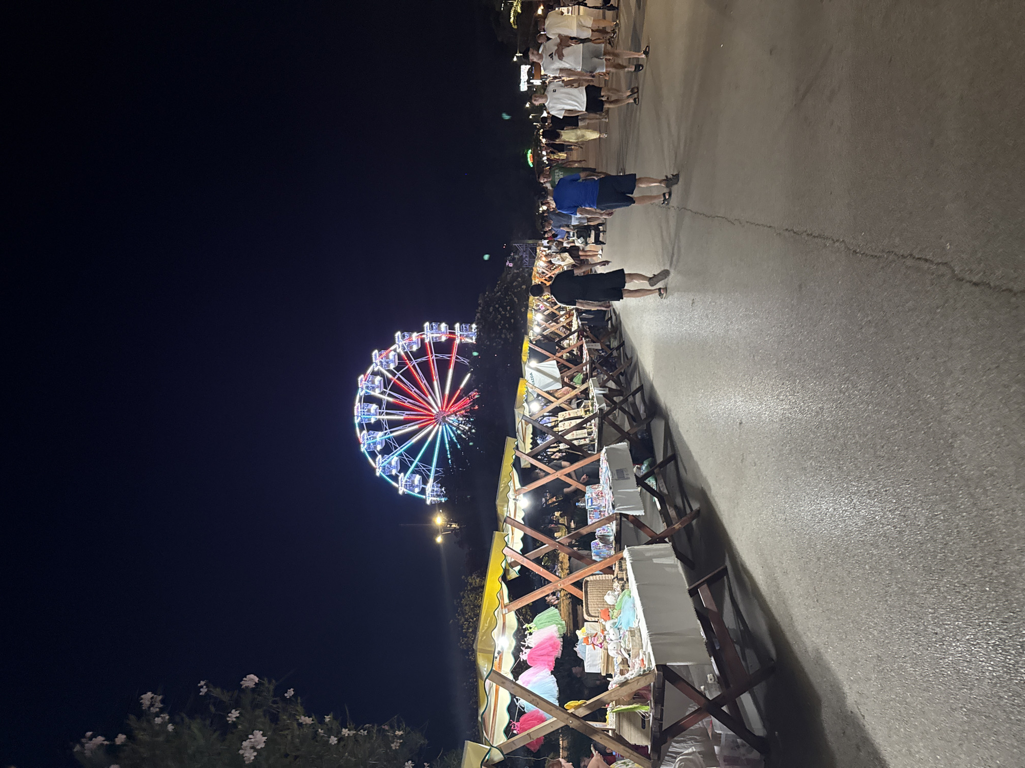 The image depicts an outdoor night market at the waterfront in Bar, Montenegro. A Ferris wheel with colorful illuminated lights is visible in the background, a prominent landmark of the city. In the foreground, a row of market stalls with yellow canopies displays various items for sale, including clothing and crafts. People are walking along the paved walkway adjacent to the market stalls, engaging in leisure activities. The scene suggests a lively atmosphere characteristic of a summer evening event.