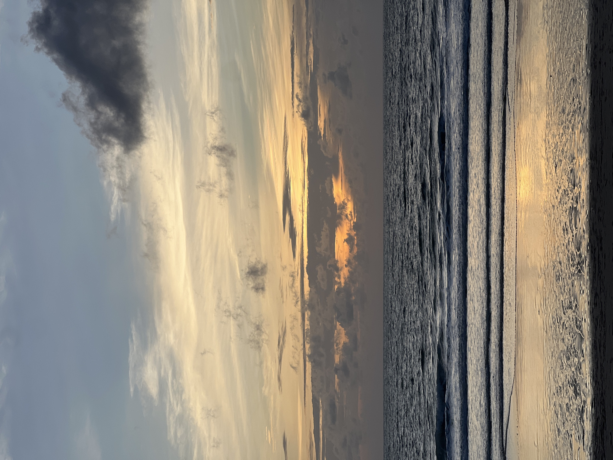 An outdoor, eye-level, full shot captures a sunset over the ocean. The sky is filled with layers of clouds in shades of blue, grey, and yellow, with the sun's rays breaking through in the distance. The ocean is a dark, rippling blue, with gentle waves lapping at the sandy shore. The wet sand reflects the colors of the sky, creating a shimmering effect. The scene is peaceful and serene, with no people or animals present. The image conveys a sense of tranquility and natural beauty. The location is the beach in Choeng Thale, Thailand.