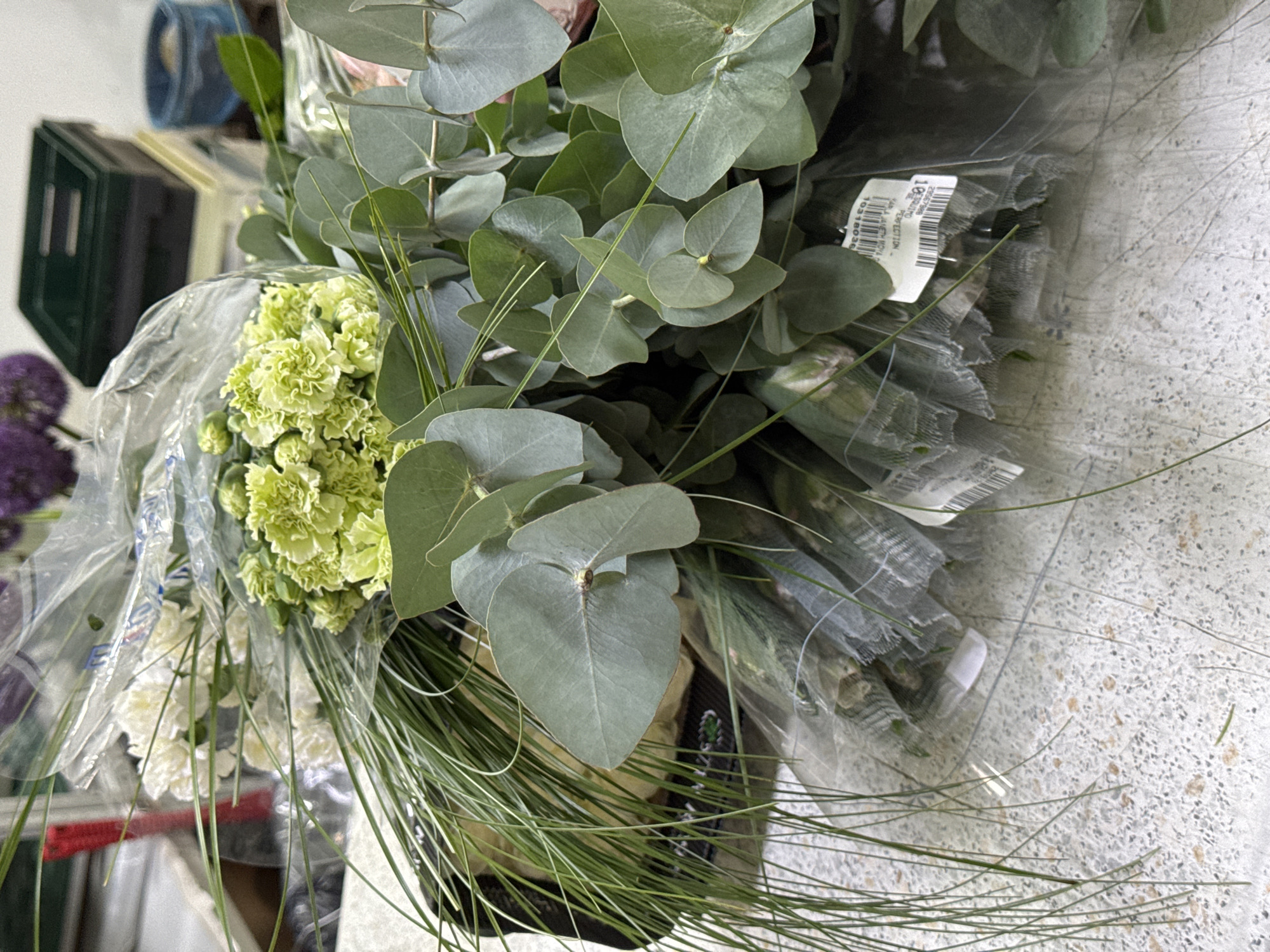 The image shows a bouquet of flowers, including eucalyptus leaves, green carnations, and white carnations, wrapped in clear plastic. Thin, green grass-like foliage is also present. Part of a price tag is visible on one of the wrapped bouquets. The backdrop shows a mottled surface, possibly a countertop or floor, and glimpses of other objects, including a green box and purple flowers. The location is unknown and not explicitly stated as Izhevsk, Russia.