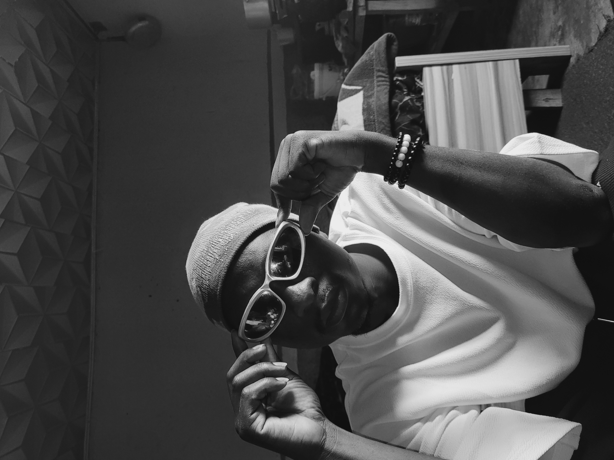 The image depicts an indoor scene in Bukuru, Nigeria, rendered in black and white. A male subject of African descent is centrally positioned, looking directly forward while adjusting white-framed, reflective sunglasses with both hands. He wears a light-colored beanie and a white, textured short-sleeved top. His left wrist displays multiple stacked bracelets composed of dark and white beads. The background features dark, plain walls and a textured ceiling with a repeating geometric pattern. To the subject's right, a partial view of a wooden or light-colored furniture piece, a white bucket, and a dark, rounded object hanging on the wall are discernible. The lighting appears to be from a single source, creating distinct shadows on the subject and in the background.