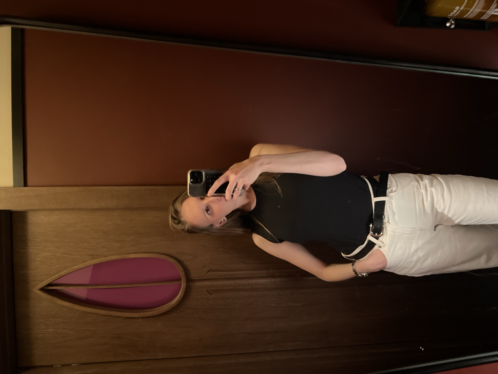 The image captures an indoor mirror selfie, likely taken in Moscow, Russia, as per the provided context. A young individual, appearing to be female, is seen holding a smartphone to capture their reflection. They are wearing a black sleeveless top, white trousers, and a black belt with a silver buckle. A silver ring is visible on their right hand, which holds the phone, and a bracelet on their left wrist.
The background of the reflection consists of a rich, deep red-brown wall in the upper section. Below this, a wooden panel or door features prominently, distinguished by a unique, elongated, leaf-shaped cutout filled with a vibrant purple or pink material. In the upper right corner of the reflected space, a black-mounted dispenser is visible, with the word "SOAP" discernable on its label. The lighting appears subdued, creating a soft, intimate ambiance. The person's face is partially obscured by the phone as they take the picture, with a neutral expression. The scene suggests a private space, possibly a bathroom or dressing area.