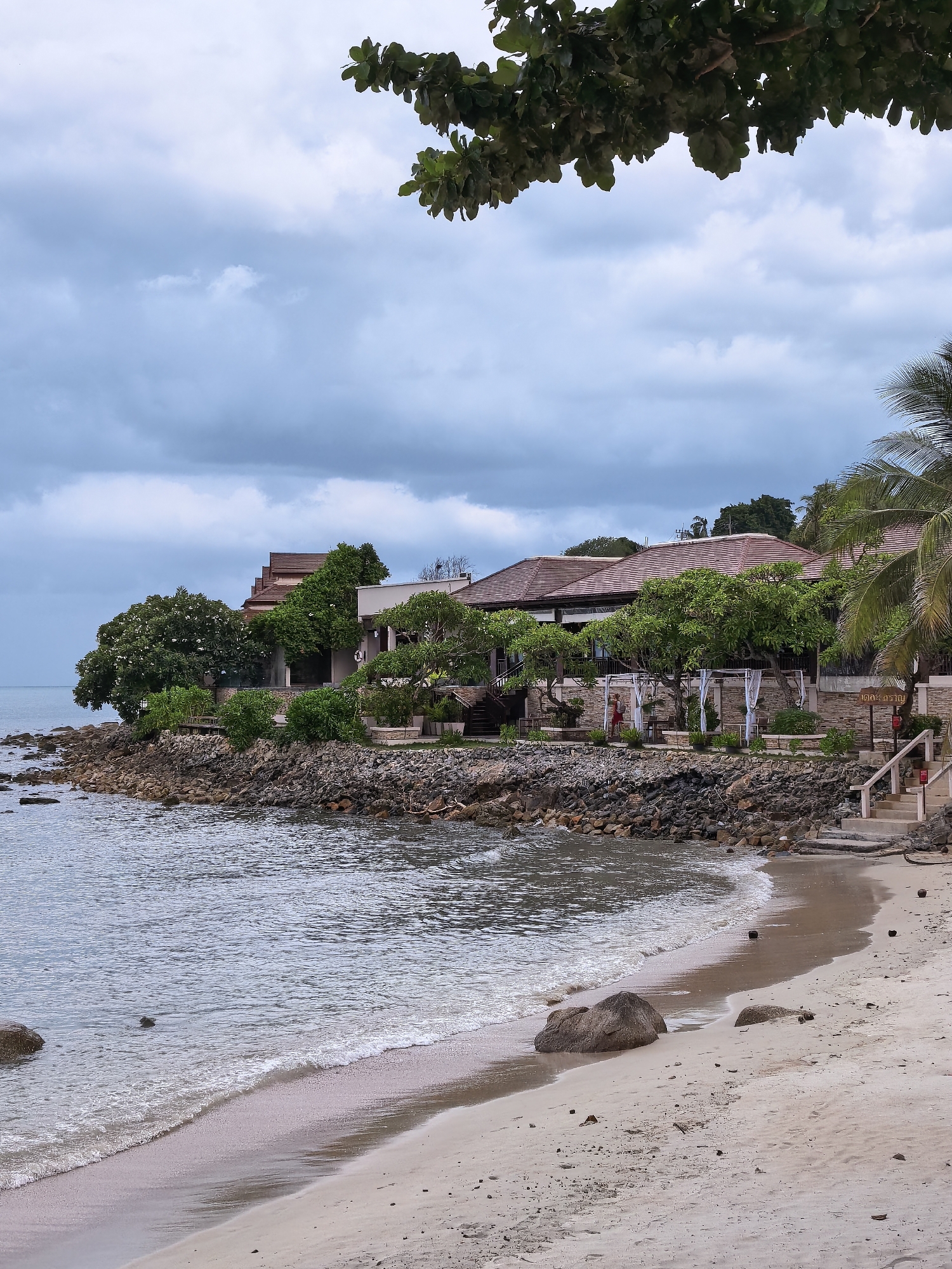 A tropical resort with traditional Thai architecture is situated on a rocky coastline in Ko Samui District, Thailand. The resort features several buildings with tiled roofs, surrounded by lush green trees and foliage. In front of the resort, a sandy beach slopes gently down to the ocean, with calm waves lapping at the shore. The sky is overcast with gray clouds, suggesting a moody or possibly impending weather change. A few large rocks are scattered on the beach and in the shallow water. The resort appears to be set up for outdoor dining or relaxation, with white draped cabanas visible. The scene evokes a sense of tranquility and natural beauty, characteristic of a beachfront resort in a tropical setting.