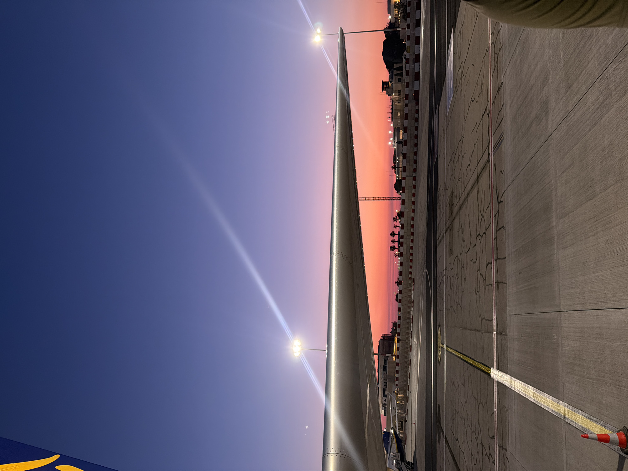 The image captures an airport scene in Luqa, Malta, at dusk. The sky transitions from deep blue to orange. The wing of a parked aircraft dominates the left side of the frame. The airport apron, marked with yellow lines, stretches into the right side of the image. Several streetlights illuminate the area and cast glares. A red and white checkered wall runs along the edge of the apron, bordering buildings and structures.