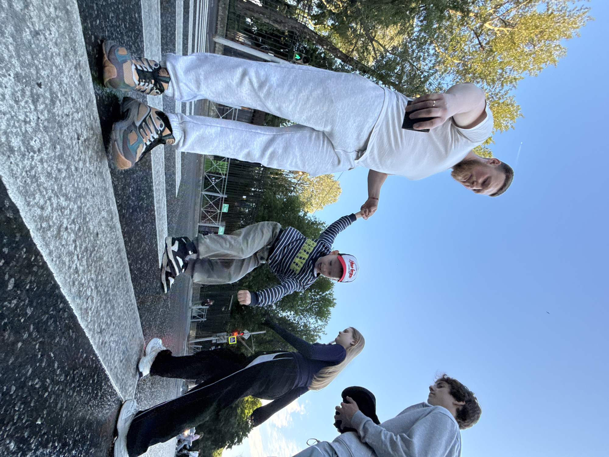 A low-angle shot captures four individuals on a pedestrian crossing at a street intersection under a clear blue sky. On the left, a man with a beard, wearing a white t-shirt and light gray sweatpants, is looking down and holding a dark object, possibly a phone, in his right hand. His left hand is outstretched. In the center, a young boy in a striped long-sleeved shirt, tan pants, and a red and white baseball cap is captured mid-jump, facing away from the viewer. He is holding the man's outstretched hand. To the right of the boy, a teenage girl with long blonde hair, wearing a dark blue long-sleeved top and black pants with white stripes, is walking away from the viewer. Further right, another young person, wearing a light gray hooded sweatshirt and dark pants, is partially visible, holding a black cap or hat in their right hand. The pedestrian crossing features white stripes on a dark asphalt road. In the background, trees with green foliage line the street, and a black metal fence is visible. Traffic signals and street signs are also present. The scene appears to be in an urban environment, potentially Perm, Russia, given the context.