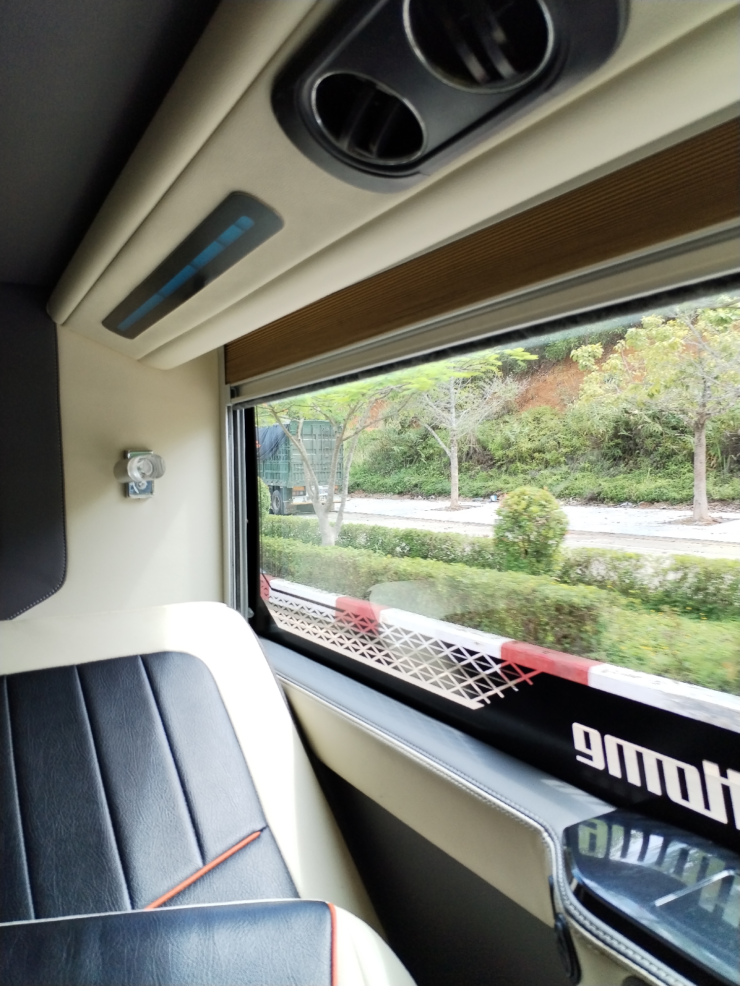 The image depicts the interior of a vehicle, likely a bus or van, in Bờ Y, Vietnam, with a view out the window. A black and white leather seat with red accents is visible in the foreground. A window displays a road with a green military truck traveling on it. The landscape outside consists of trees, bushes, and an embankment. A utility pole is also apparent. The window has a red and white border at its bottom. The ceiling of the vehicle features air conditioning vents and lighting.