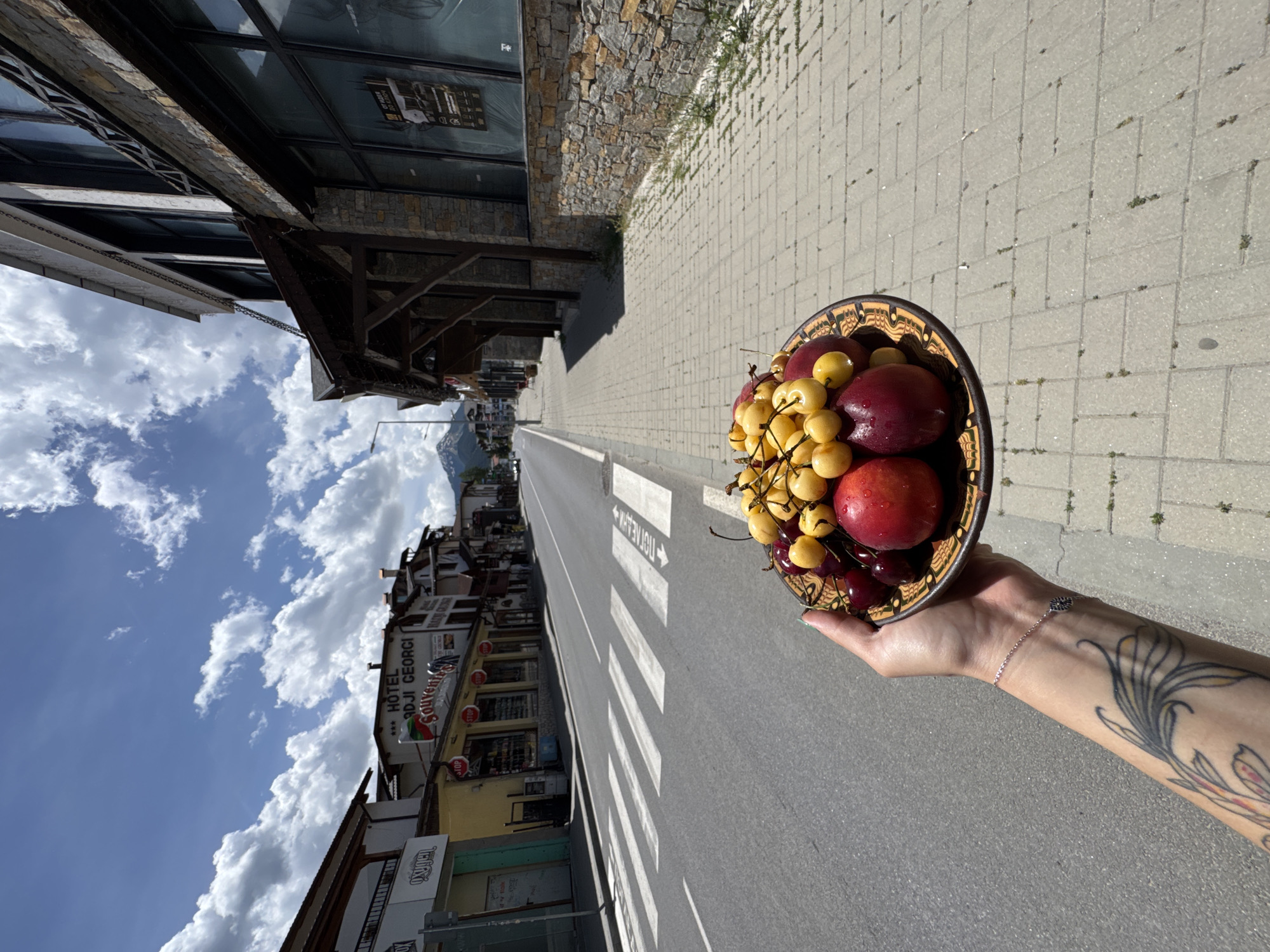 A low-angle, medium shot captures a hand holding a decorative ceramic plate filled with ripe cherries and peaches on a sidewalk in Bansko, Bulgaria. The plate, adorned with an intricate brown and orange pattern, is positioned in the foreground, obscuring the lower part of the hand and arm, which bears a tattoo. The sidewalk, constructed of light gray interlocking bricks, stretches into the background. To the left, a paved road with a white pedestrian crossing runs alongside a row of buildings. The buildings, featuring wooden balconies and stone facades, house shops with signs such as "HOTEL ADJI GEORGI" and "Souvenirs." A mountain range, partially visible in the distance, is silhouetted against a bright blue sky dotted with white, fluffy clouds. The sun is shining, casting shadows and illuminating the scene.