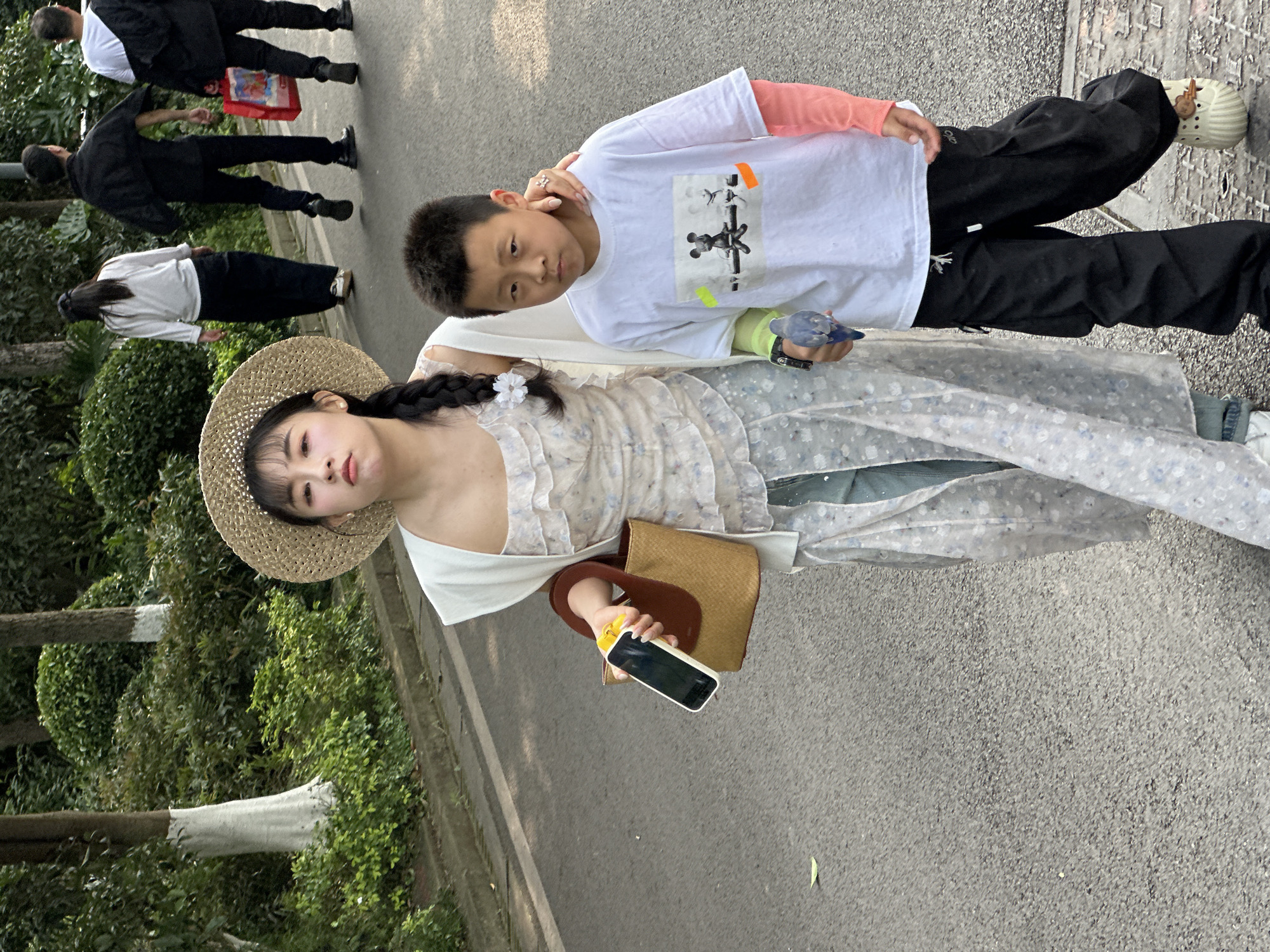 The image shows a woman and a boy standing on a paved road. The woman wears a straw hat, a light-colored dress, and carries a brown bag and a phone. The boy wears a white t-shirt and black pants. They are in a park-like setting with trees and other people walking in the background. The location is Liangjiang, China.