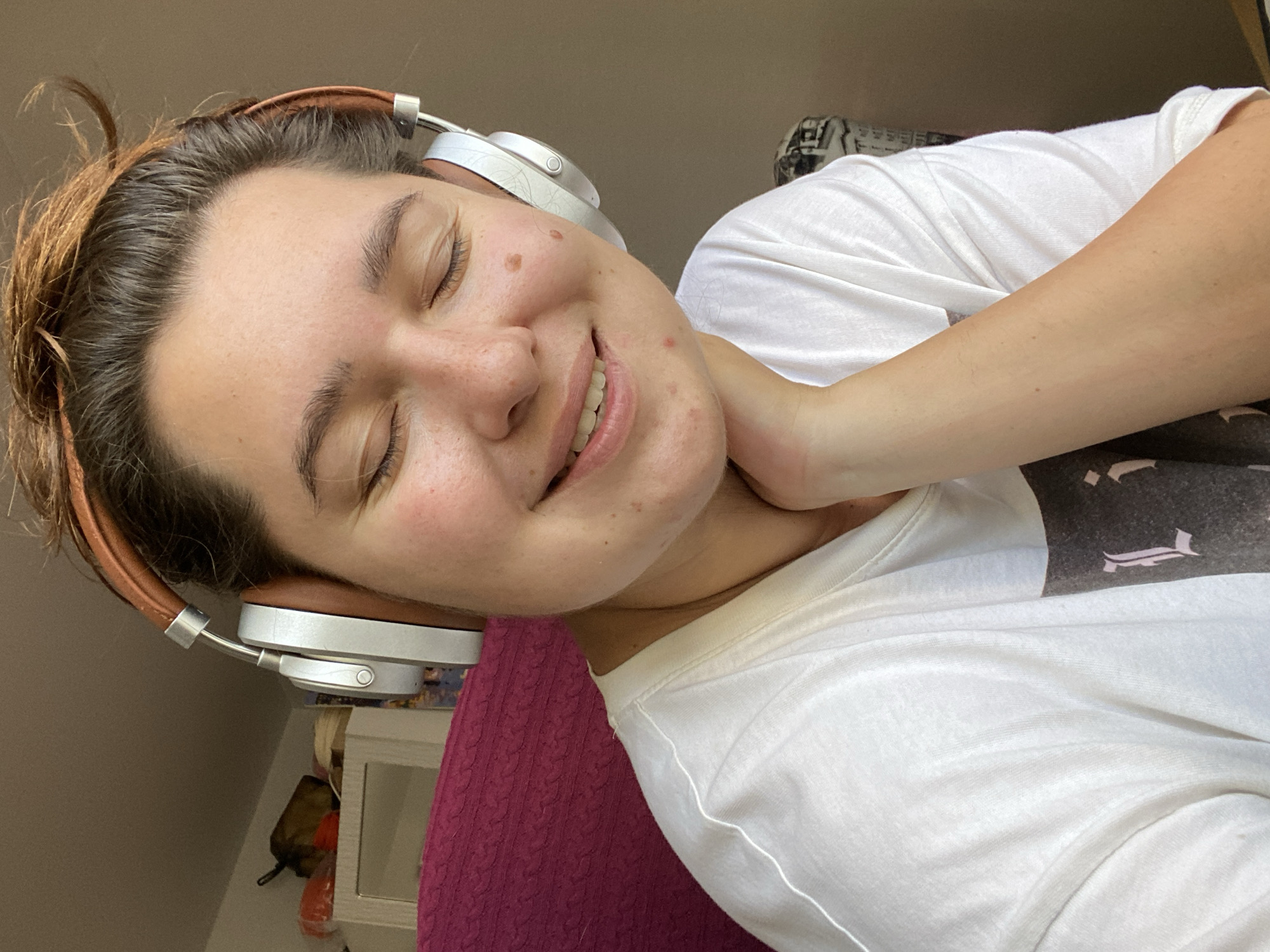 A young woman is shown wearing over-ear headphones with a brown leather headband and silver earcups. She has her eyes closed and is smiling with her chin resting on her hand. She is wearing a white t-shirt. The background appears to be indoors with a hint of furniture and a pink textured surface. There is no indication of the location in Perm, Russia.