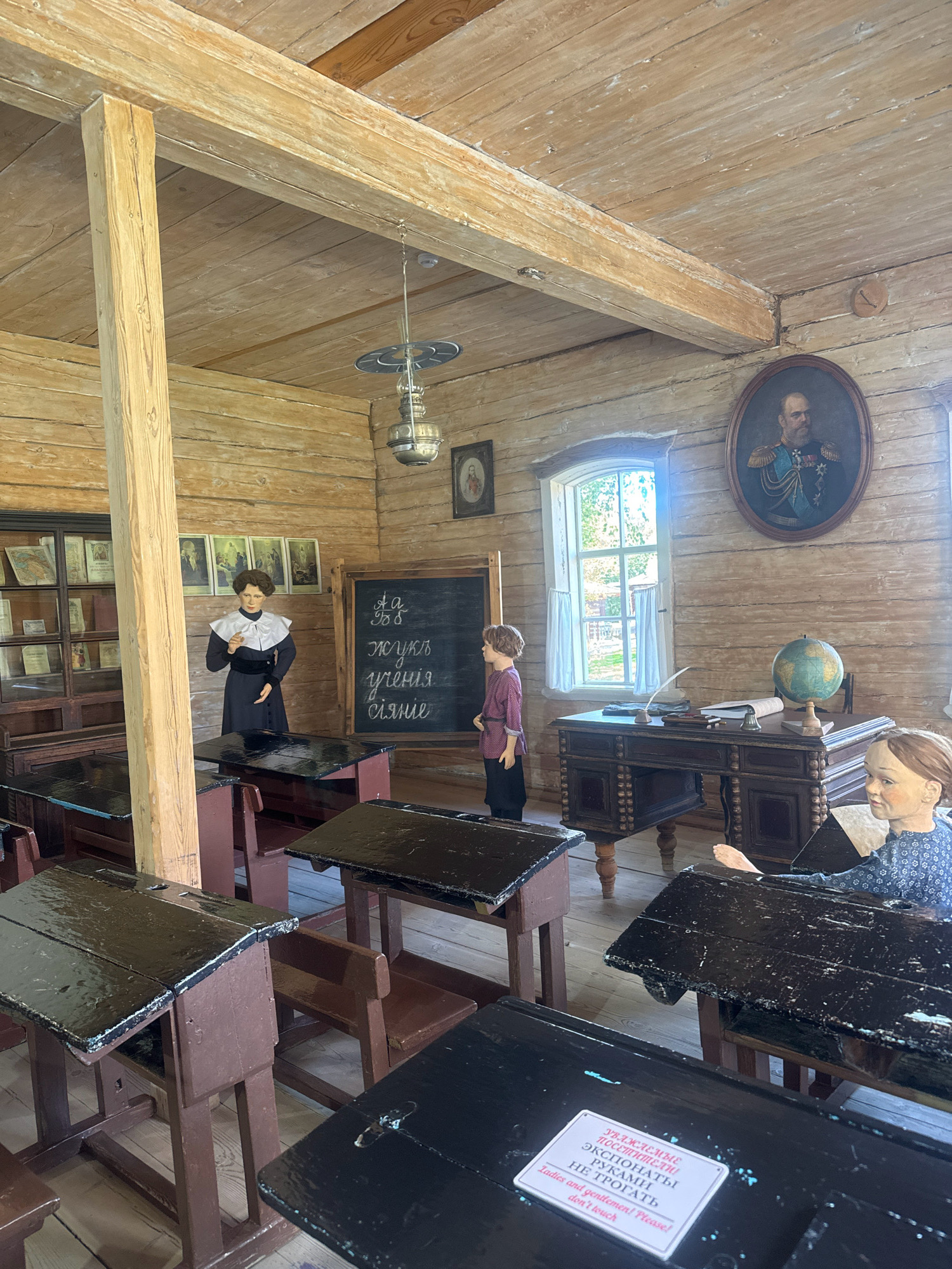 The image depicts a classroom interior at the Taltsy Museum (Irkutsk, Russia). The room is constructed from logs, with a wooden ceiling and floor. In the foreground, multiple antique wooden desks with black, glossy tops are arranged in rows. A mannequin of a woman, dressed in a dark dress and a white collar, stands near a blackboard with Cyrillic writing. To her right, a young boy mannequin is partially visible. A teacher's desk is positioned by a window, featuring a globe, a quill pen, and an inkwell. A framed portrait of a man in military uniform hangs on the wall. Another mannequin, a young girl, is seated at a desk in the middle ground. A sign with text in both Russian and English, reading "Ladies and gentlemen! Please don't touch," is affixed to one of the desks. The overall scene portrays a historical classroom setting.