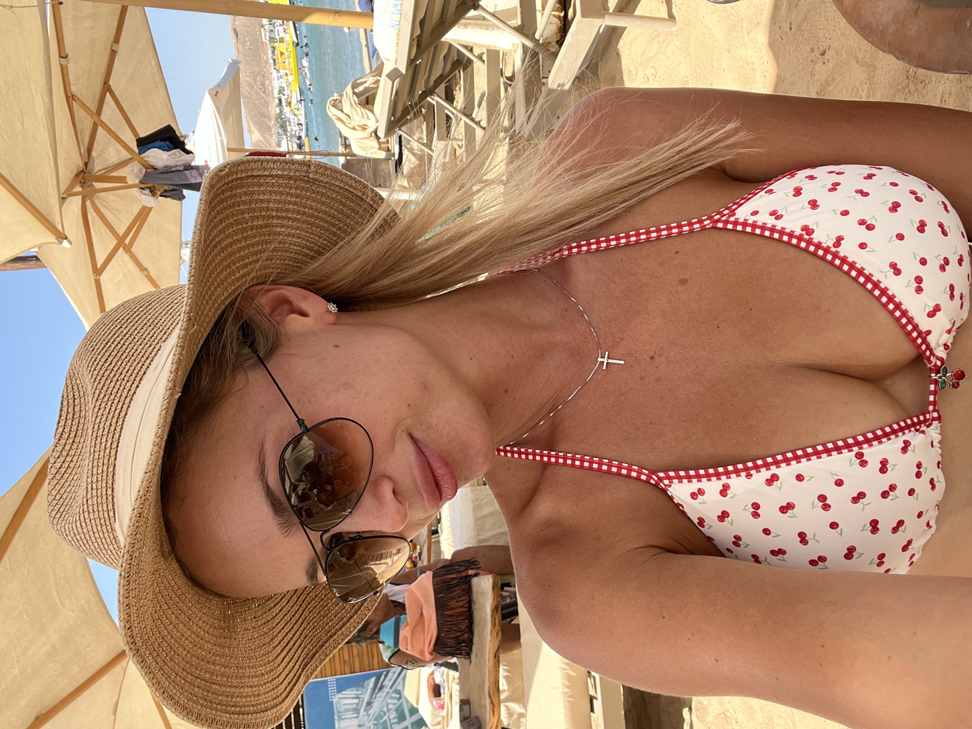 A blonde woman wearing a straw hat and sunglasses poses for a selfie on a beach in Sharm El Sheikh, Egypt. She is wearing a white bikini with a red cherry pattern and a red and white checkered trim. A silver necklace with a cross pendant hangs around her neck. The background shows beach chairs, umbrellas, and the sea with boats and buildings in the distance. The sky is clear and blue.
