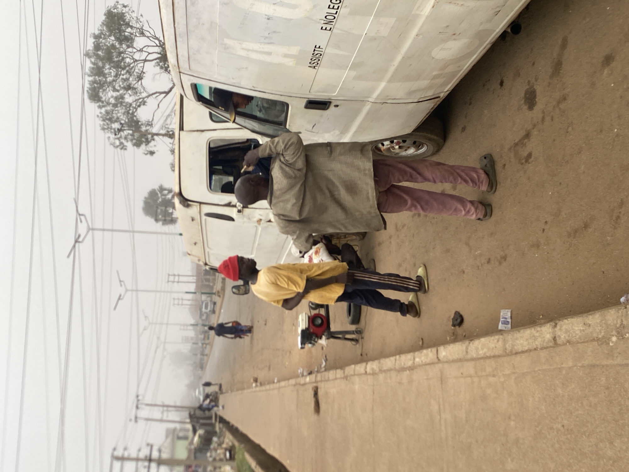 On a road in Bukuru, Nigeria, under a hazy sky, a light-colored van is parked. A man in a gray top and maroon pants stands by the driver's side door, appearing to interact with the driver. Another man, wearing a yellow shirt, dark pants with stripes, and a red cap, stands nearby. Between them sits a small generator. The surrounding environment includes power lines, trees, and a sidewalk. Additional vehicles and pedestrians are visible further down the road.