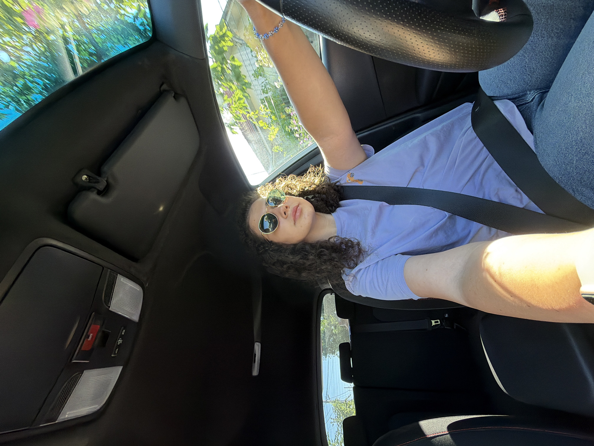 A young woman with curly dark hair and sunglasses is driving a car on a sunny day. She is wearing a light purple t-shirt and blue jeans, with a seatbelt fastened across her chest. The interior of the car is visible, including the steering wheel, dashboard with an SOS button, and rear seats. The exterior environment shows trees and houses through the windows, suggesting an urban or suburban setting. The image is captured from inside the car, looking forward and slightly to the right. The presence of an SOS button in the car suggests it's likely a modern vehicle. The architecture and lush greenery visible through the windows are consistent with areas in Sofia, Bulgaria.
