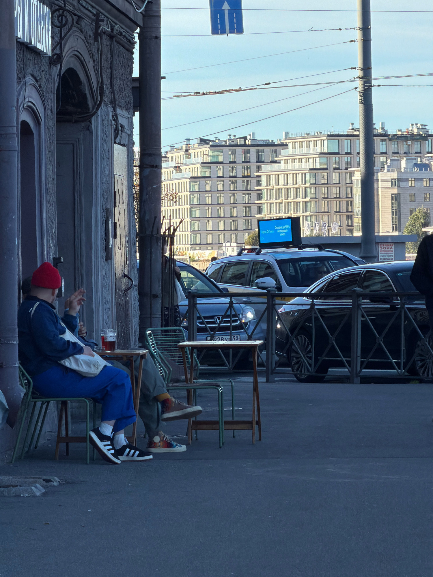 In Saint Petersburg, Russia, on a sunny day, two people are seated at a small table outside a building with a textured gray facade. The person on the left wears a red beanie, a blue jacket, and blue pants. They are holding a cigarette and appear to be smoking. A white bag is on their lap. The person on the right wears a darker colored jacket and pants, with one leg bent. A glass of amber liquid, possibly beer, sits on the table. Two simple metal chairs and two small metal tables are present. In the background, modern apartment buildings with many windows dominate the skyline under a clear blue sky. Several cars are visible on the street, including a black sedan and a silver SUV with a digital advertisement on its roof. Electrical wires crisscross the sky.