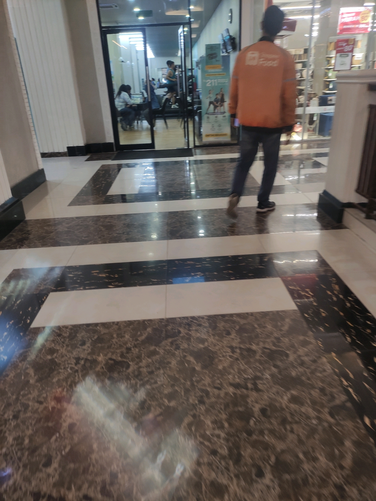 The image depicts a wide shot of a modern shopping mall interior. The floor is tiled in a geometric pattern of black, white, and dark brown marble-like squares and rectangles. In the background, a glass-enclosed gym area is visible, with several people exercising on treadmills and other equipment. One person is on a treadmill, wearing a blue top and black shorts. Another person is sitting at a table with a laptop. The foreground shows the legs and lower torso of a person wearing dark jeans and black sneakers, walking away from the camera. This person is wearing an orange jacket with a white logo on the back, which appears to be for a food delivery service. The overall ambiance suggests a busy commercial area. This scene is located in Sinduadi, Indonesia.