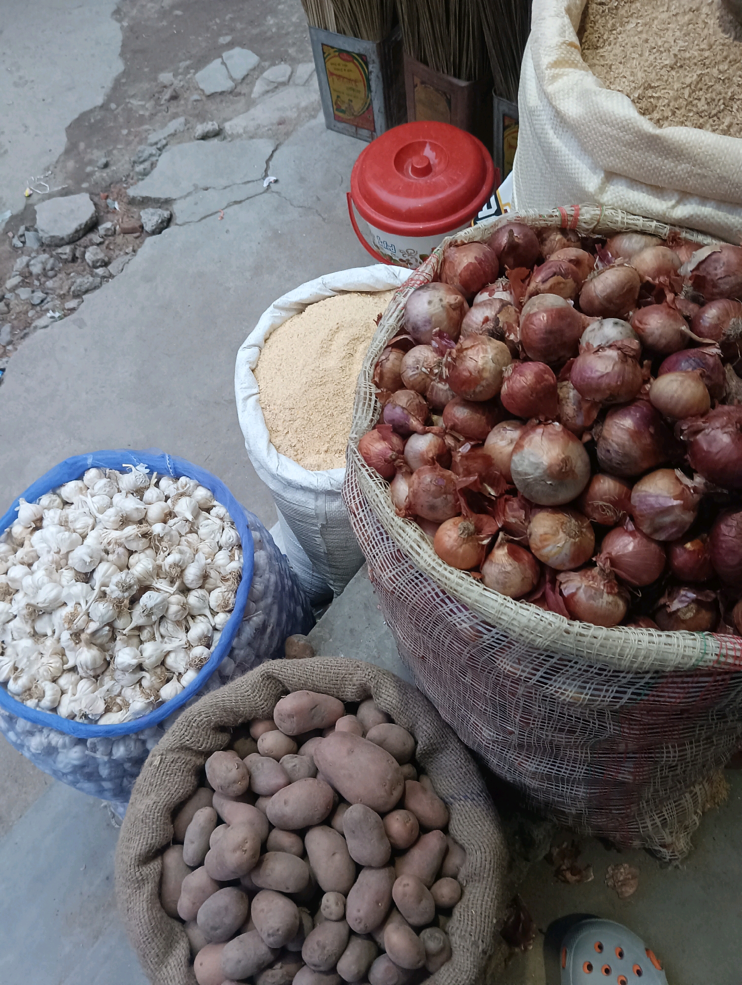 A top-down, medium shot captures a street-level market scene in Dakhin Parsa, India. The foreground features a burlap sack overflowing with potatoes, their earthy brown skins varying in color and texture. To the left, a large blue mesh bag is filled with pale white garlic bulbs, their papery skins visible. Behind the garlic, a large white woven bag contains a pile of light yellow grain, possibly rice or lentils. Dominating the right side of the frame is a large net bag brimming with red onions, their dry skins showing shades of red and brown. A red plastic bucket with a lid sits to the left of the onions, near a framed sign with text in an Indian script and a broom behind it. The ground is a mix of cracked concrete and rough, uneven patches. The overall scene suggests a bustling marketplace with agricultural produce on display for sale.