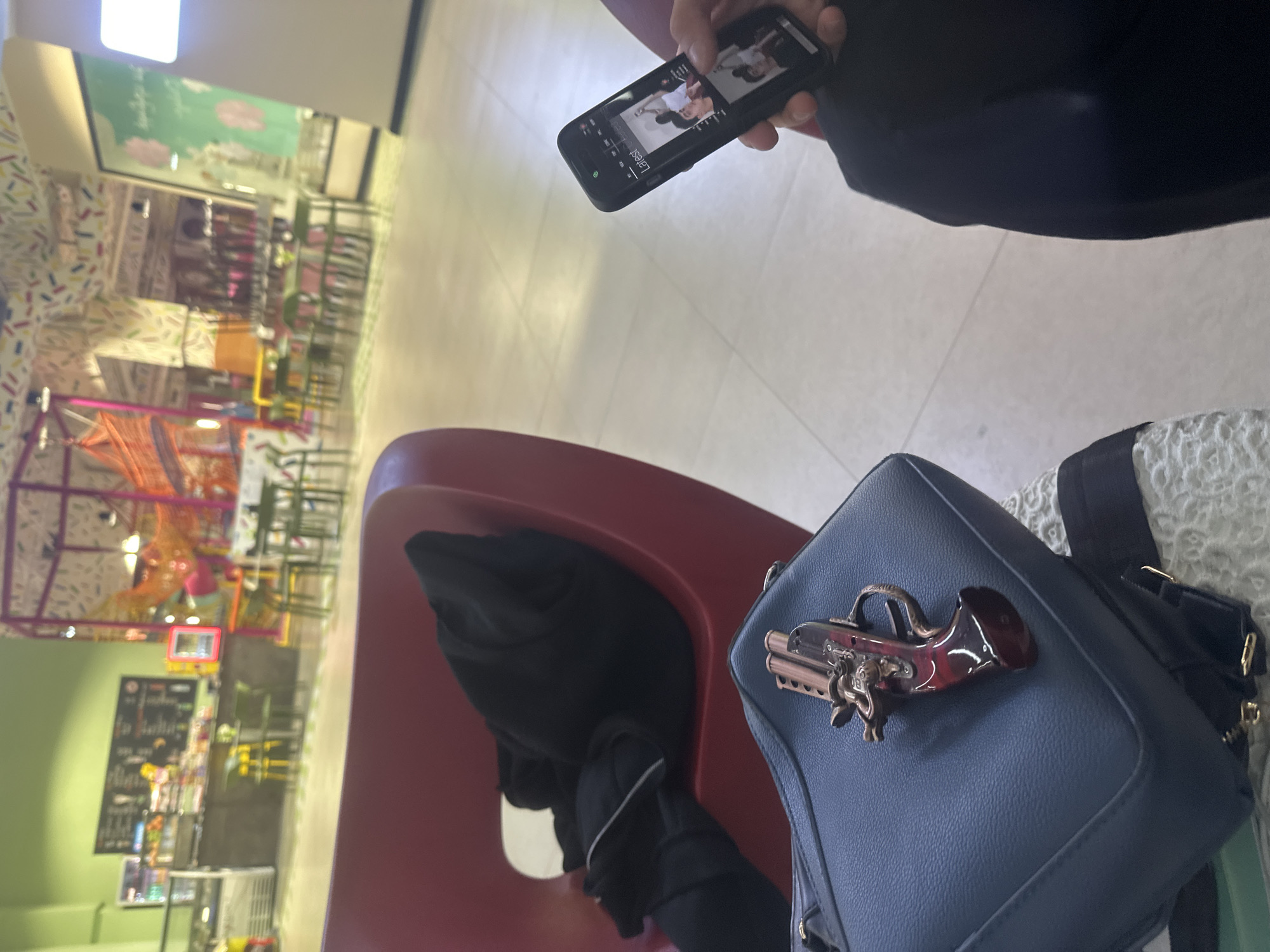 The image appears to be taken indoors, possibly in a shopping center or public area with retail spaces. In the foreground, there's a person holding a smartphone, presumably taking a picture. A blue purse with a gun-shaped ornament on it is visible, resting on a surface alongside other personal belongings, including black gloves.
The background includes elements suggesting a commercial environment, possibly a food court or a children's play area, with tables and chairs visible in the distance. The overall lighting is bright, indicating daytime or well-lit indoor conditions.