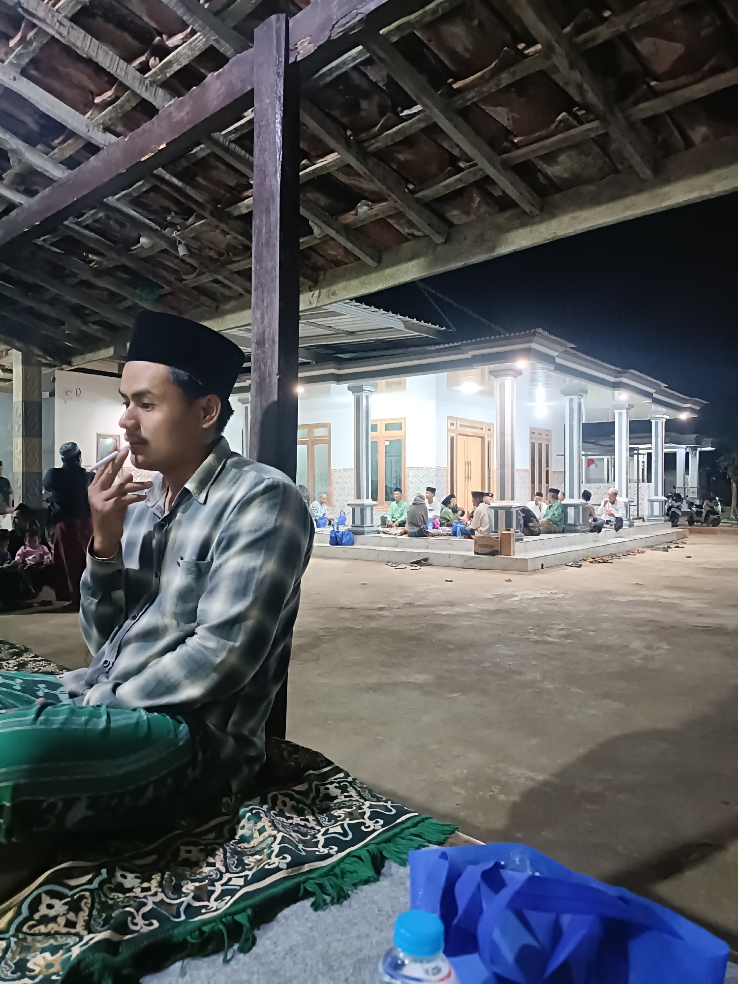 The image depicts an outdoor gathering at night in the city of Bicorong, Indonesia, likely a social or religious event. In the foreground, a young man wearing a black peci (cap) and a checkered shirt is seated, smoking a cigarette. He is on a patterned mat with green fringe, with a blue bag and a water bottle visible nearby. Behind him, other people are seen sitting and talking in front of a well-lit building with a covered porch supported by white pillars. The building appears to be a mosque or a community center. The scene is lit by artificial lights, creating a warm and ambient atmosphere. The overall environment suggests a relaxed and communal gathering.