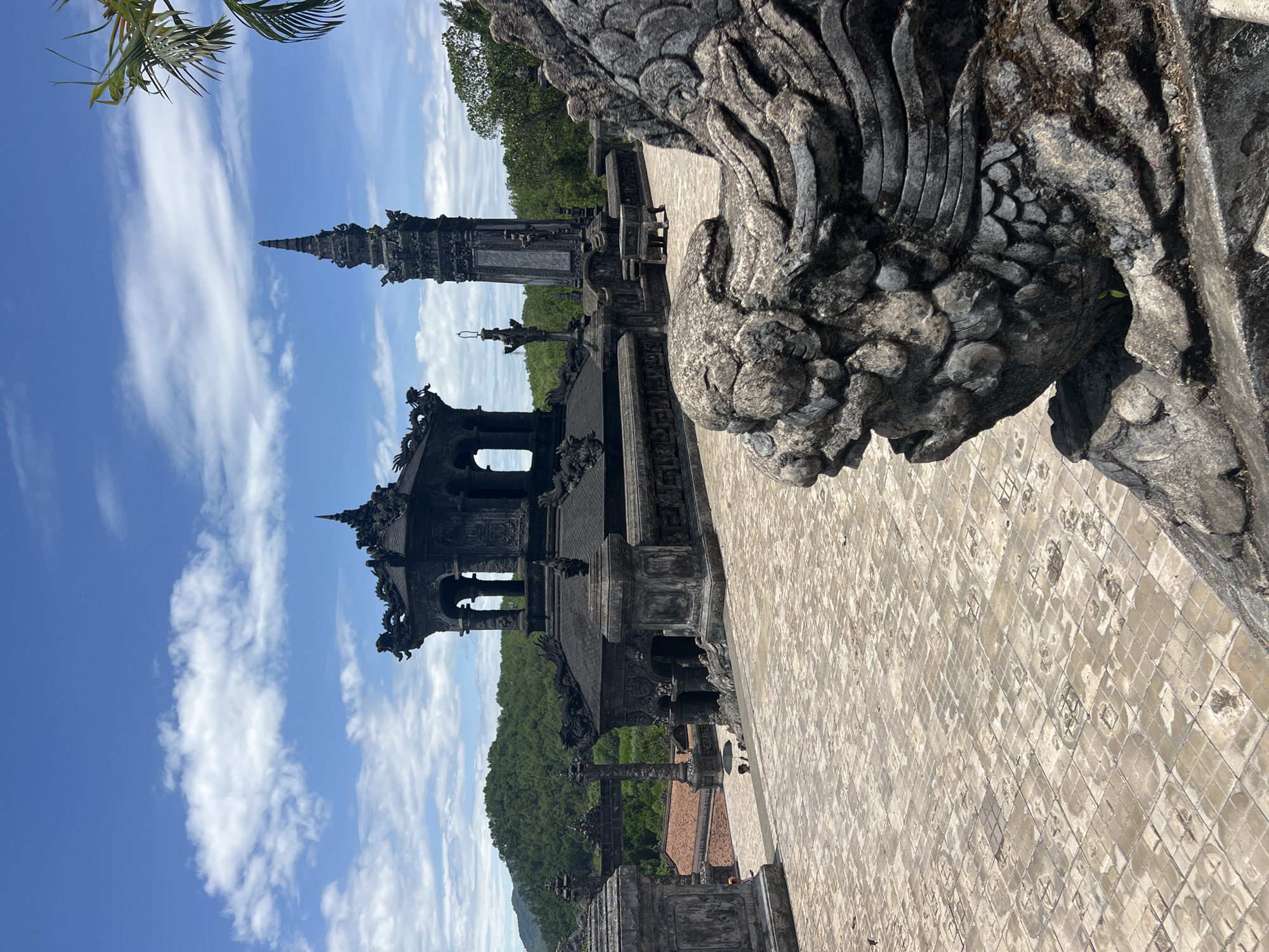 The image depicts the courtyard of the Tomb of Khai Dinh in Thuy Xuan Ward, Vietnam. The foreground features a weathered, stone dragon carving, its textured scales and weathered surface indicative of age and exposure to the elements. Behind the dragon, a vast, tiled courtyard stretches out, the tiles arranged in a grid pattern and showing signs of wear and discoloration.
Dominating the midground are several ornate, dark stone structures characteristic of Vietnamese imperial architecture. To the left, a multi-tiered pavilion with intricate carvings and dragon motifs stands against a backdrop of lush green hills. To the right, a tall, slender tower or stupa rises towards the sky, also adorned with detailed ornamentation. In the distance, more architectural elements can be seen, including a large archway and tiled roofs.
The sky above is a vibrant blue, dotted with fluffy white clouds. A palm frond extends into the frame from the upper right corner, suggesting a tropical or sub-tropical climate. Two individuals are visible in the middle ground, one holding an umbrella, suggesting a sunny day. The overall scene conveys a sense of historical grandeur and the enduring beauty of ancient architecture set within a natural landscape.