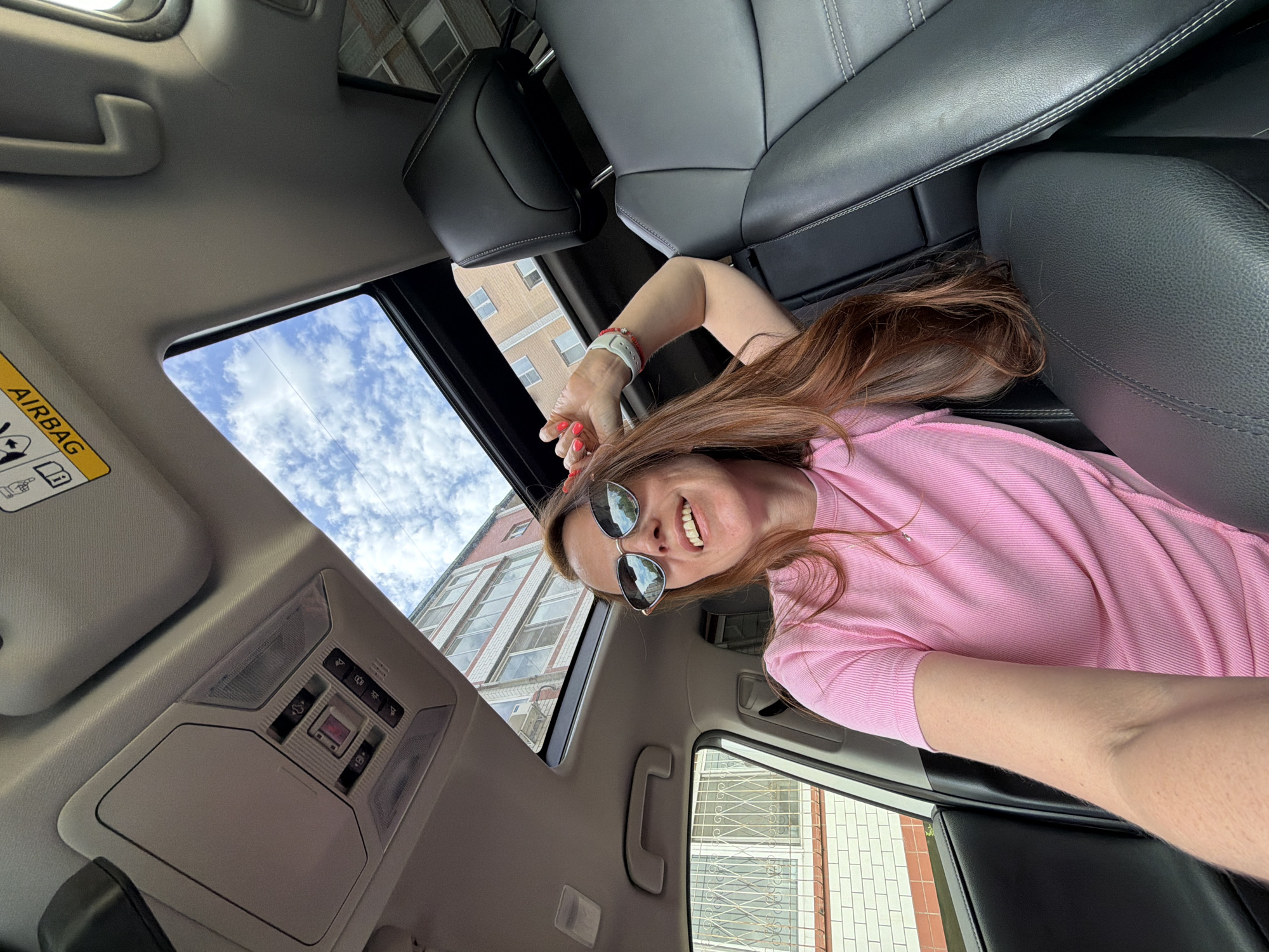 A young woman with long auburn hair and sunglasses is taking a selfie inside a car. She is wearing a pink t-shirt and a white smartwatch. The car has a sunroof open, revealing a cloudy blue sky and buildings outside. The interior of the car is dark leather. The image appears to have been taken on a sunny day in Birobidzhan, Russia.