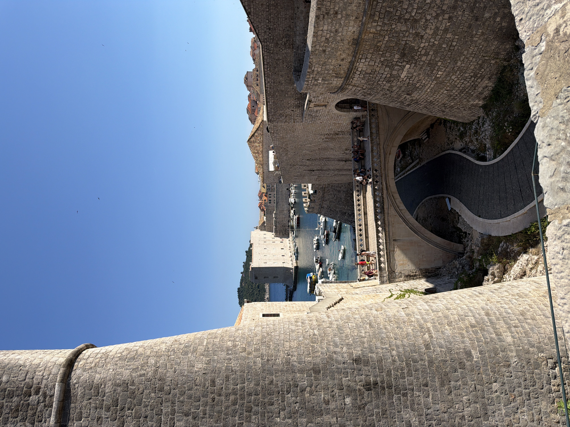 The image captures a view from the city walls of Dubrovnik, Croatia, looking down towards the harbor. The sky is a clear, bright blue, indicating a sunny day, likely in the late morning or early afternoon given the strong sunlight casting shadows. The ancient stone walls, constructed from rough-hewn blocks, dominate the foreground and midground, showcasing their impressive scale and texture.
Below the walls, a picturesque harbor is visible, filled with numerous small boats and yachts moored in the clear blue water. In the distance, more buildings with terracotta roofs, characteristic of Dubrovnik's Old Town, are nestled against a backdrop of lush green hills. A curved, paved pathway with railings snakes down from the wall, and several people can be seen walking on it or sitting on the steps, enjoying the view.
The scene is one of historical grandeur and vibrant activity. The sturdy stone fortifications suggest the city's rich past, while the bustling harbor and the presence of visitors point to a popular tourist destination. The atmosphere appears peaceful and serene, with the sunlight illuminating the ancient architecture and the sparkling water. No visible text is present in the image.